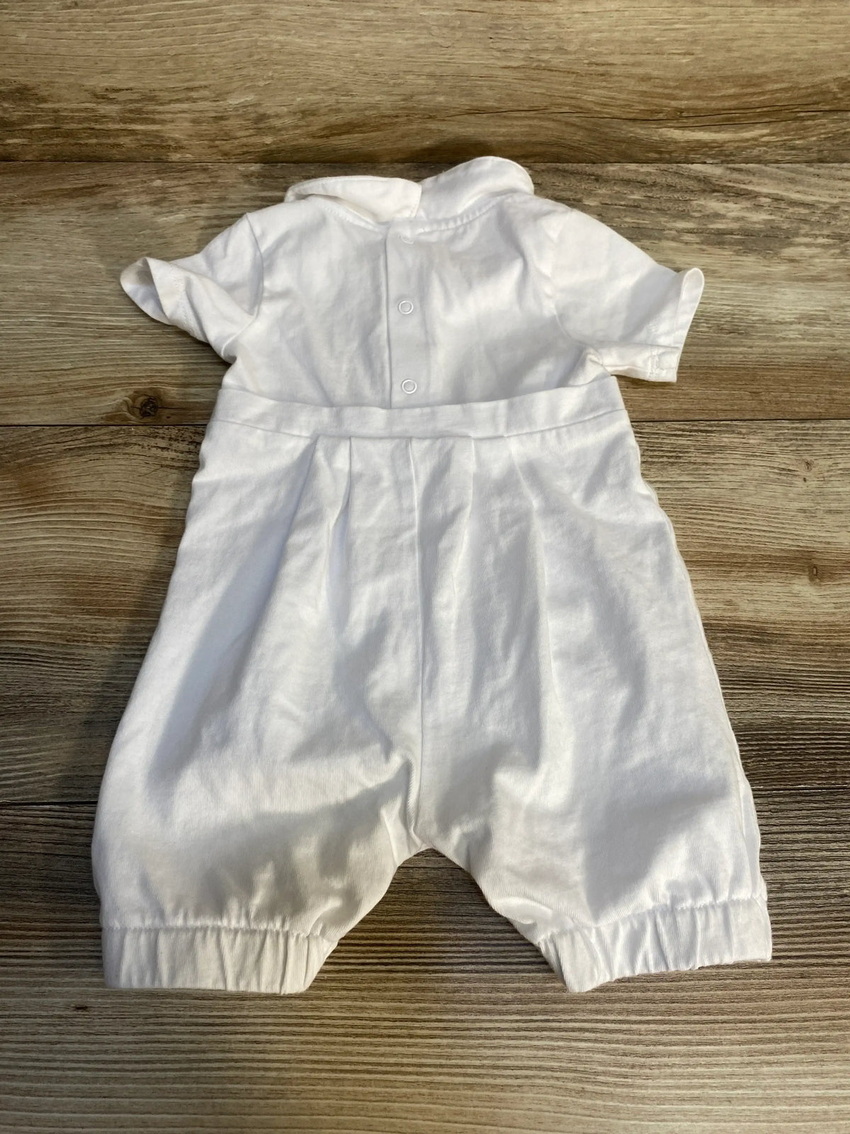 Front view of white christening romper showcasing short sleeves, pleated waist, and snap closures on a wooden surface