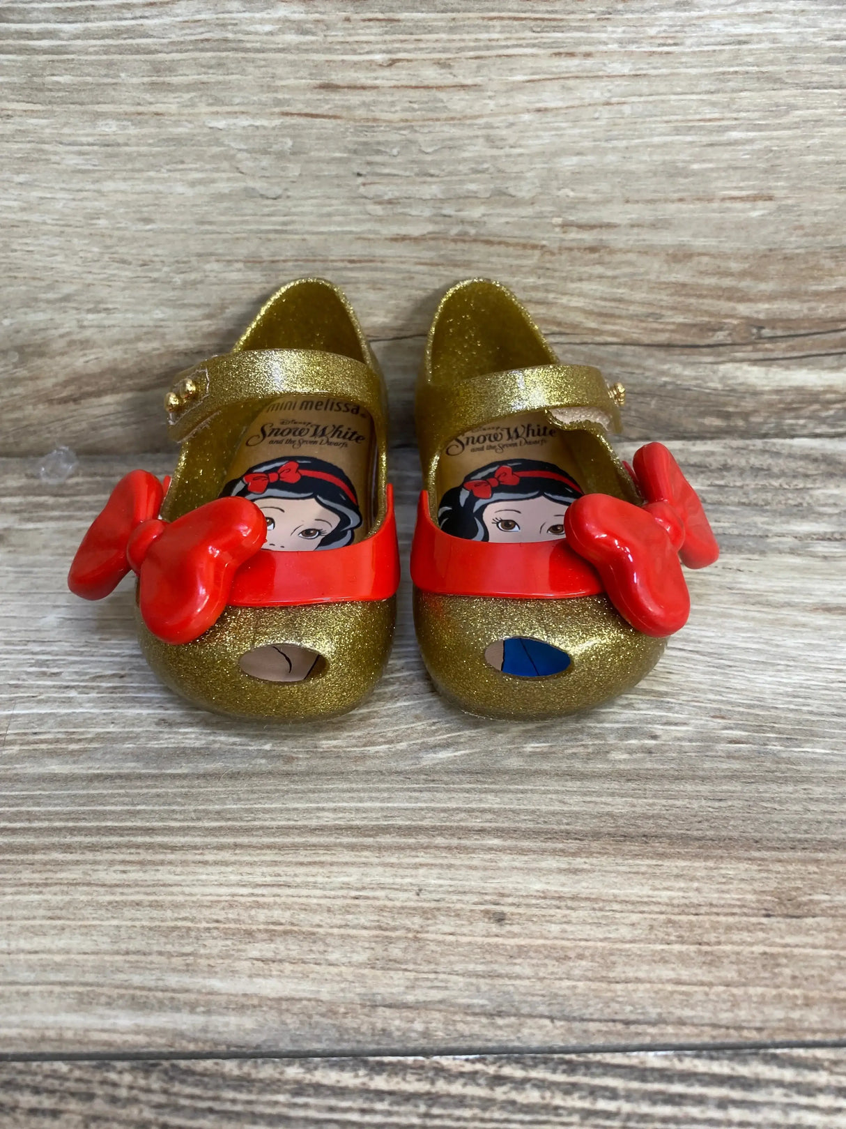 Front view of Mini Melissa Ultragirl shoes showcasing gold glitter, red bows, and Snow White insoles on wood background