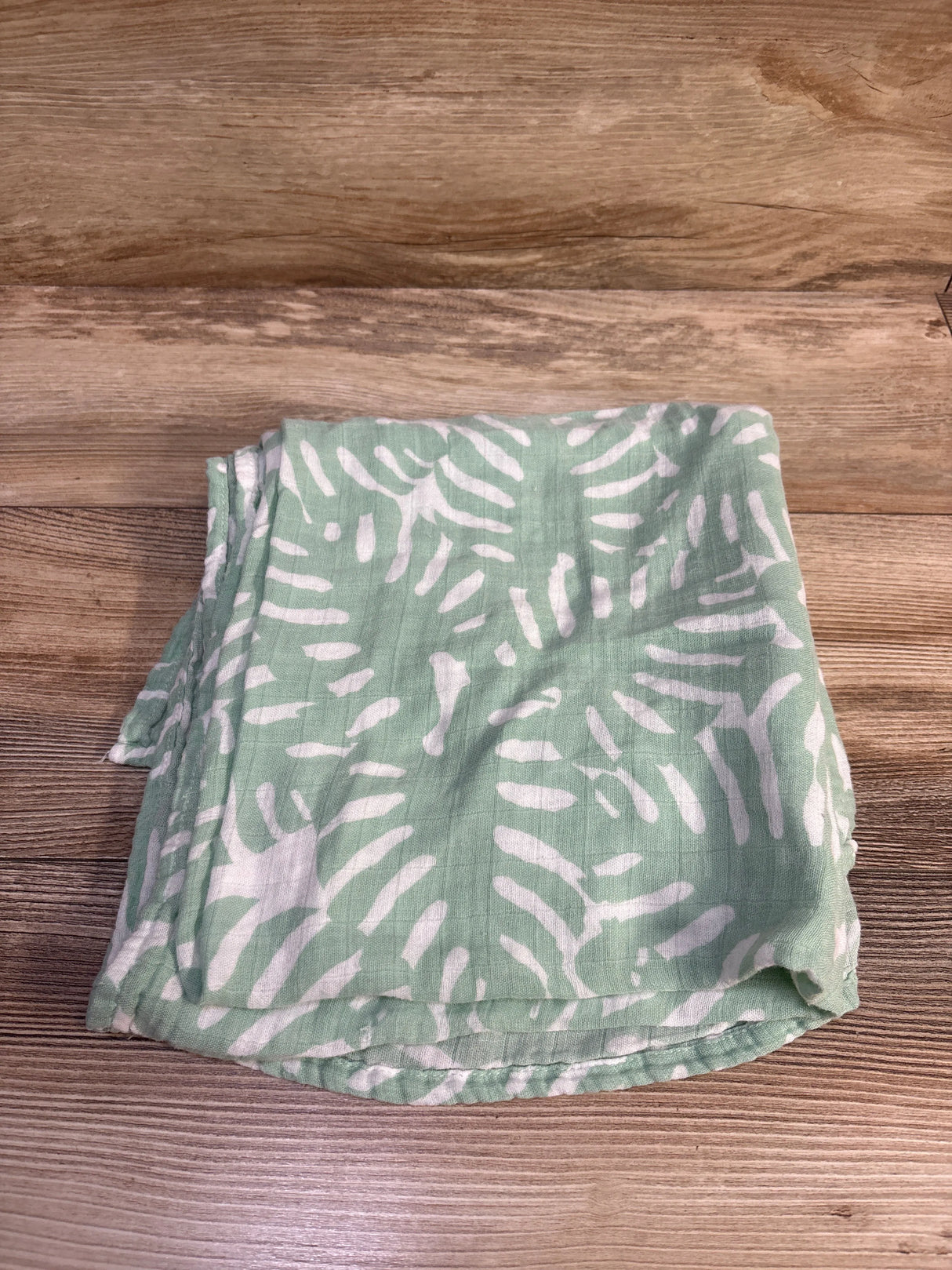 Top view of folded Momcozy bamboo swaddle blanket showing green fabric with white abstract leaf pattern on wood background