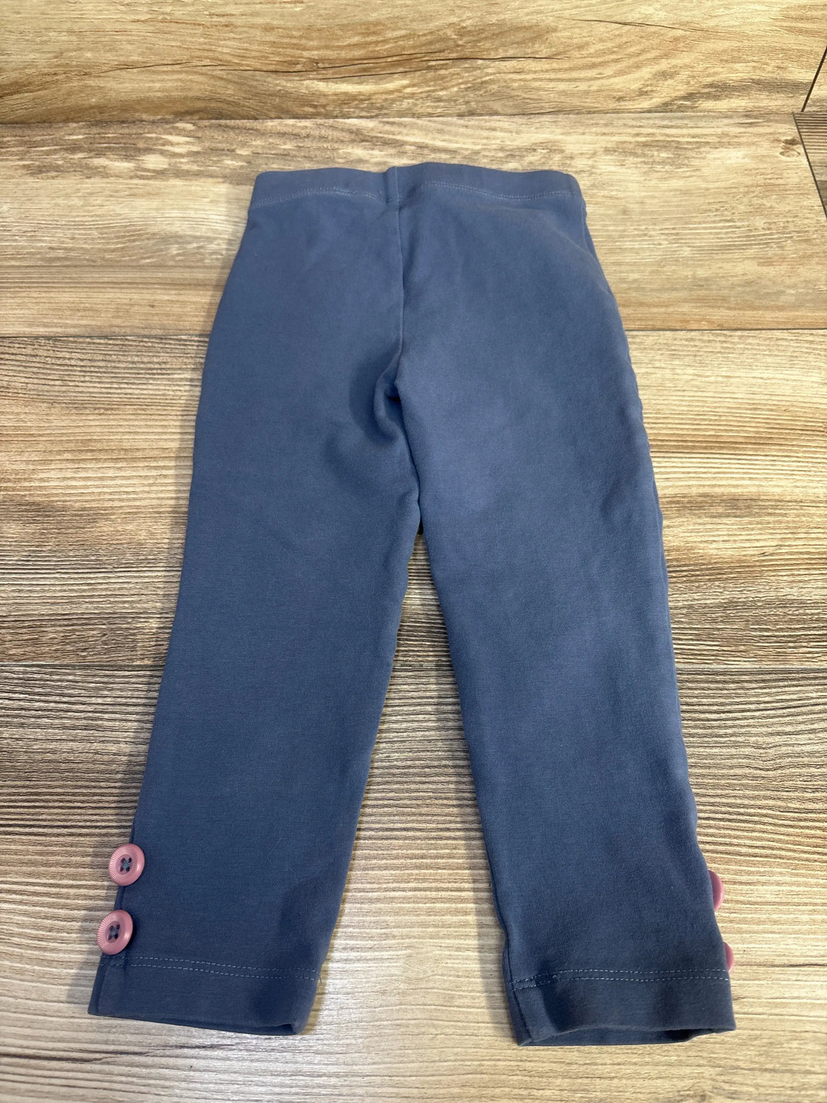 Top-down flat lay of toddler navy blue leggings size 2T showing two pink buttons at ankle on wood background