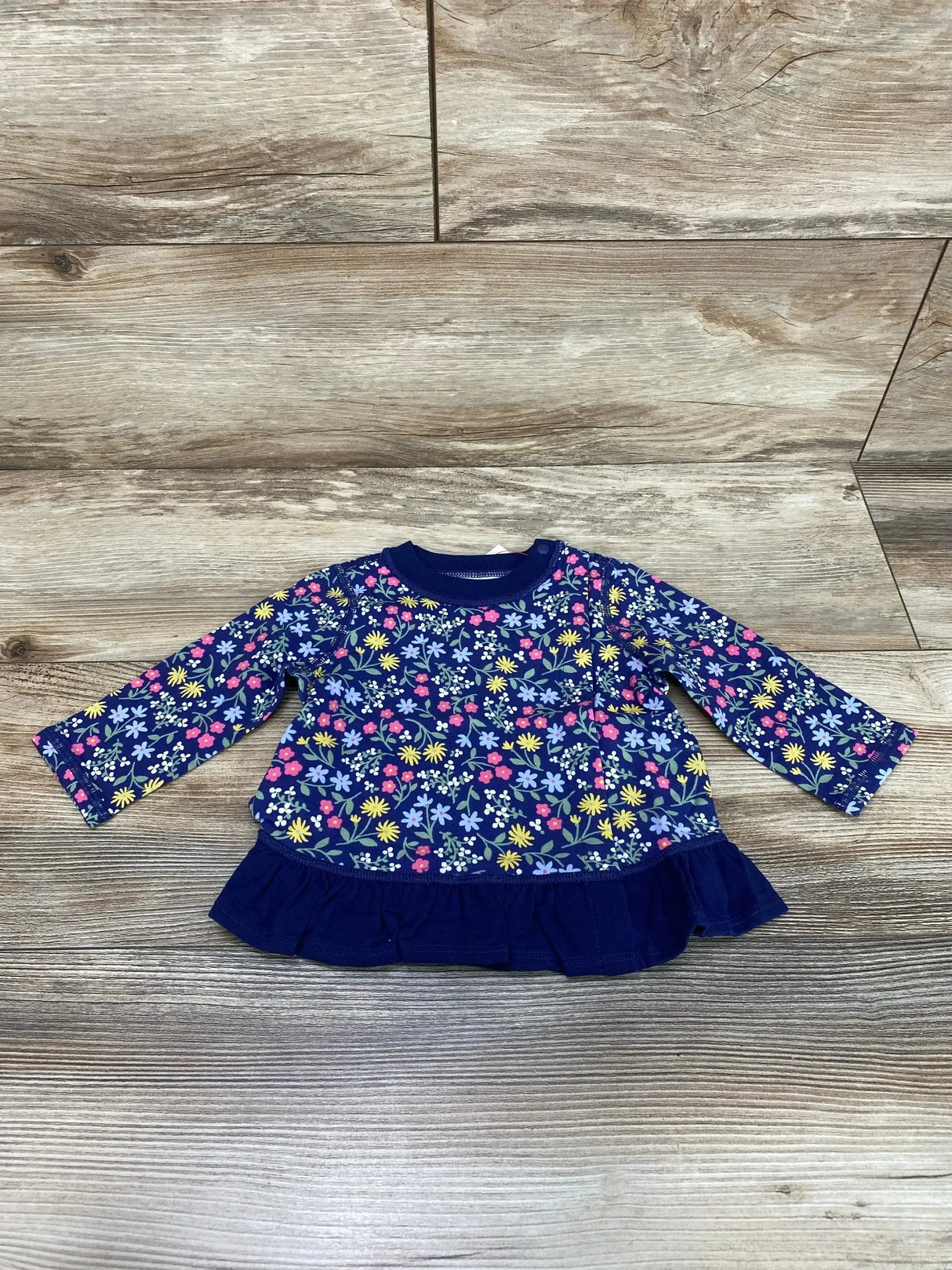 Flat lay of Cat and Jack navy peplum baby top with floral print, long sleeves, ruffled hem and shoulder snaps on wood floor