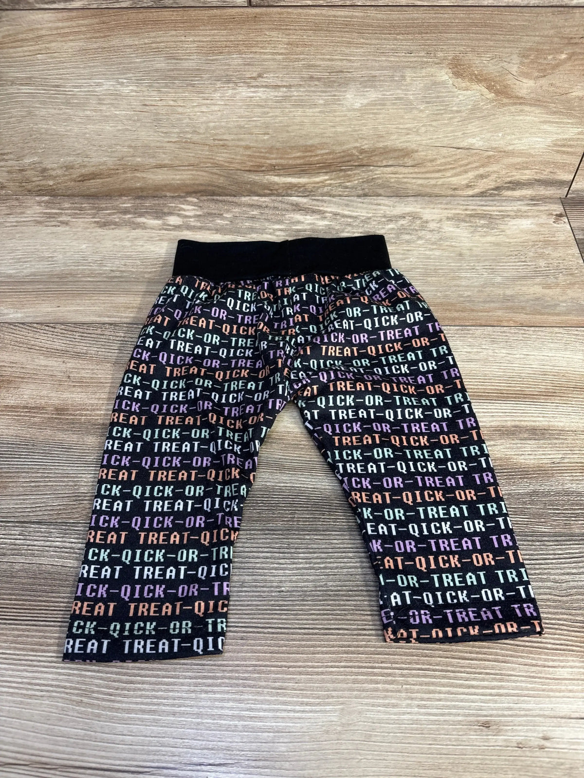Front view of black Trick Or Treat pants with multicolor all over text print and elastic waistband on a wood surface