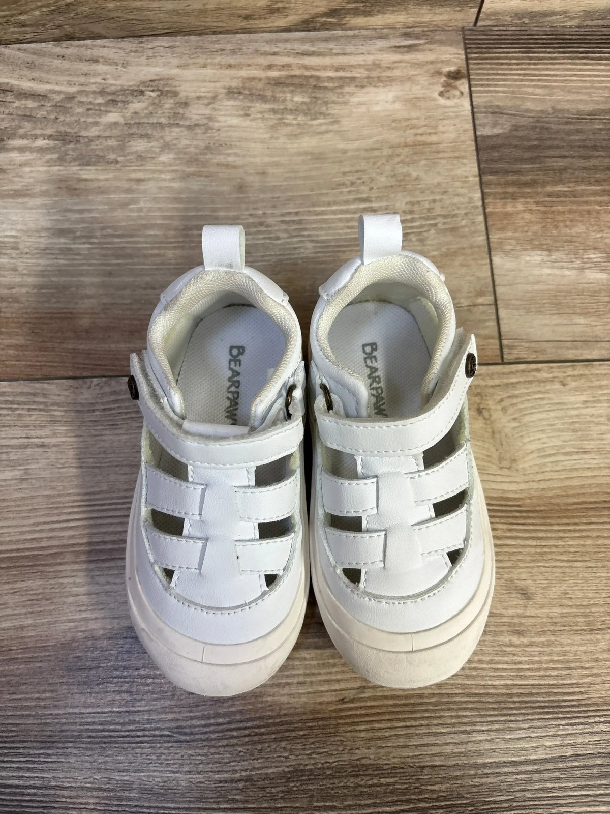 Top down view of Bearpaw white kids fisherman sandals with woven closed toe straps and Velcro ankle strap on wood floor