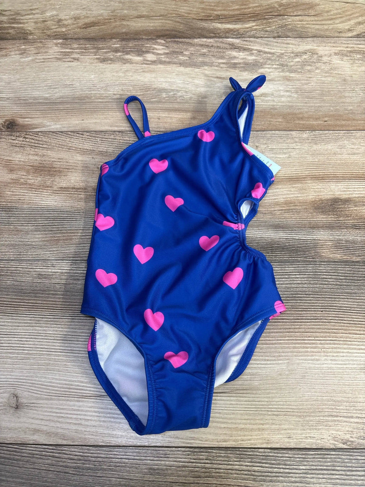 Front view of Cat and Jack blue one piece cut out swimsuit with pink heart pattern and tie shoulder straps on a wood grain background