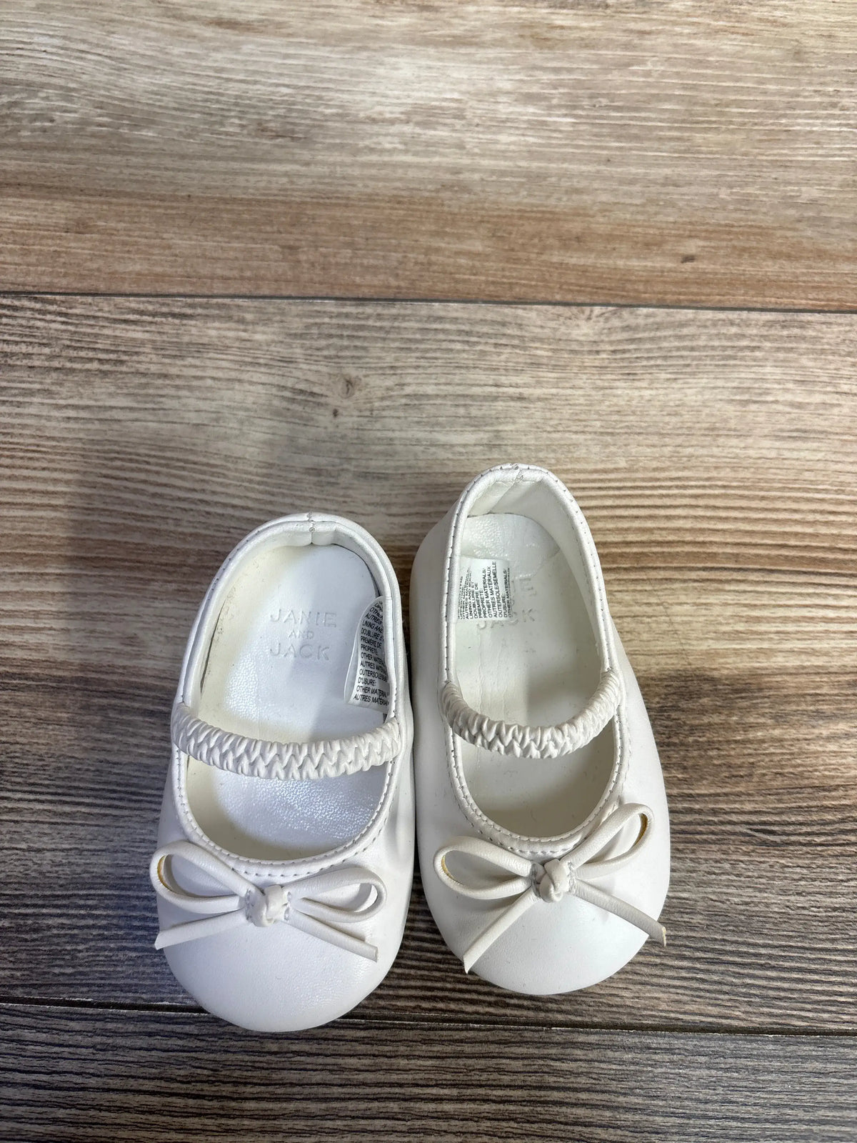 Pair of Me n Mommy To Be white soft sole ballet flats with bow details on a wood background