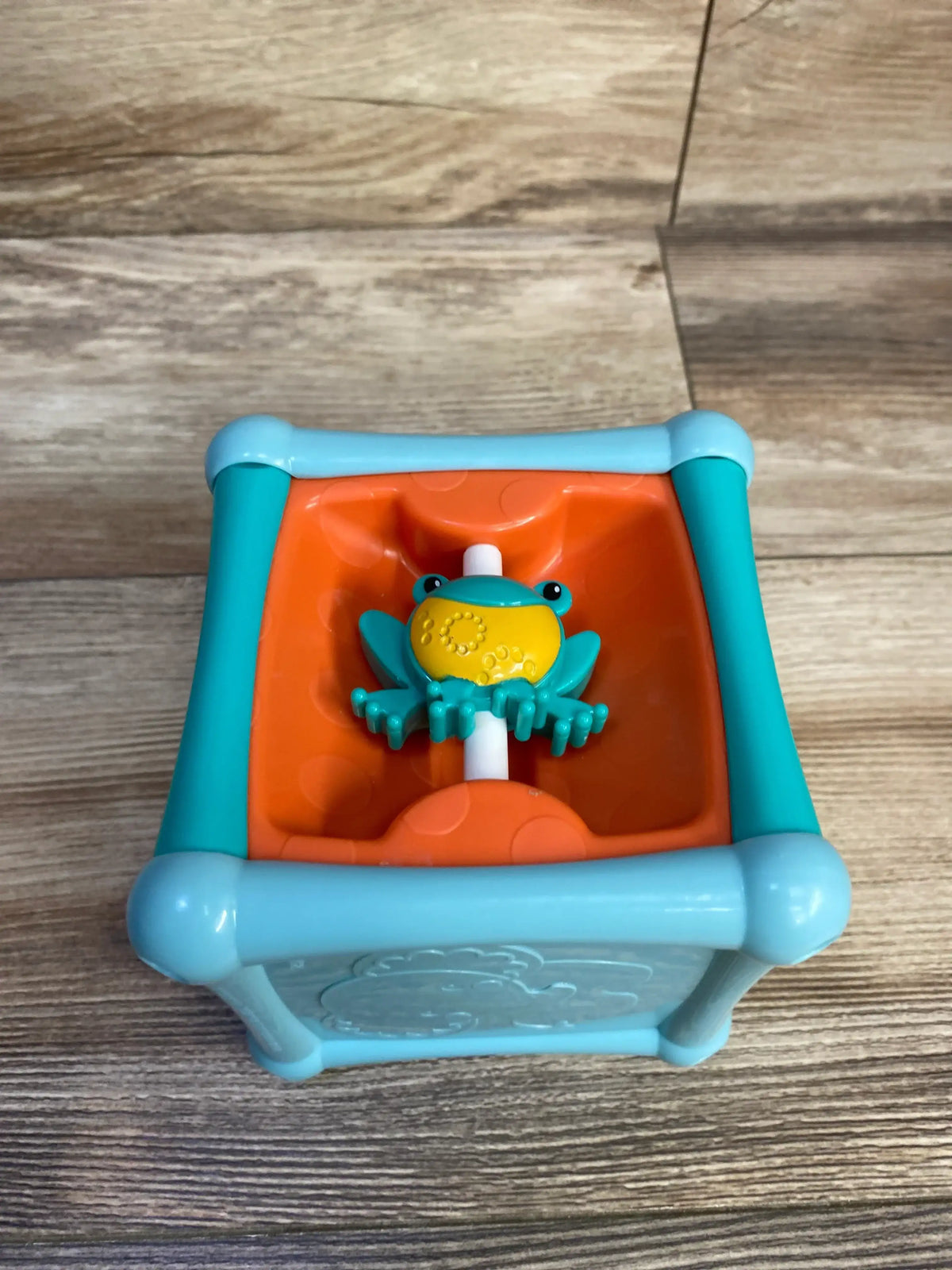 Top view of Fisher Price activity cube showing turquoise frog bead on white spindle inside orange cup and light blue frame
