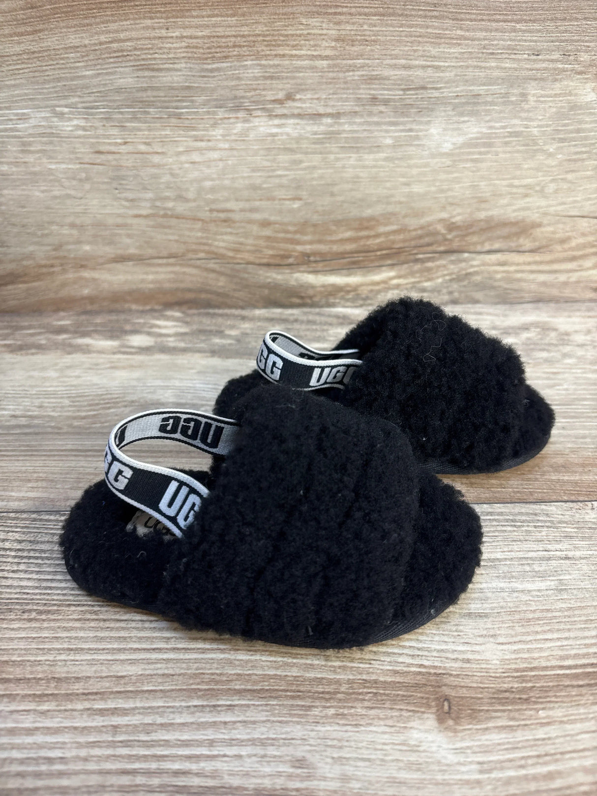 Angled view of UGG Fluff Yeah toddler slides in black showing fuzzy upper and elastic UGG heel strap on wood background