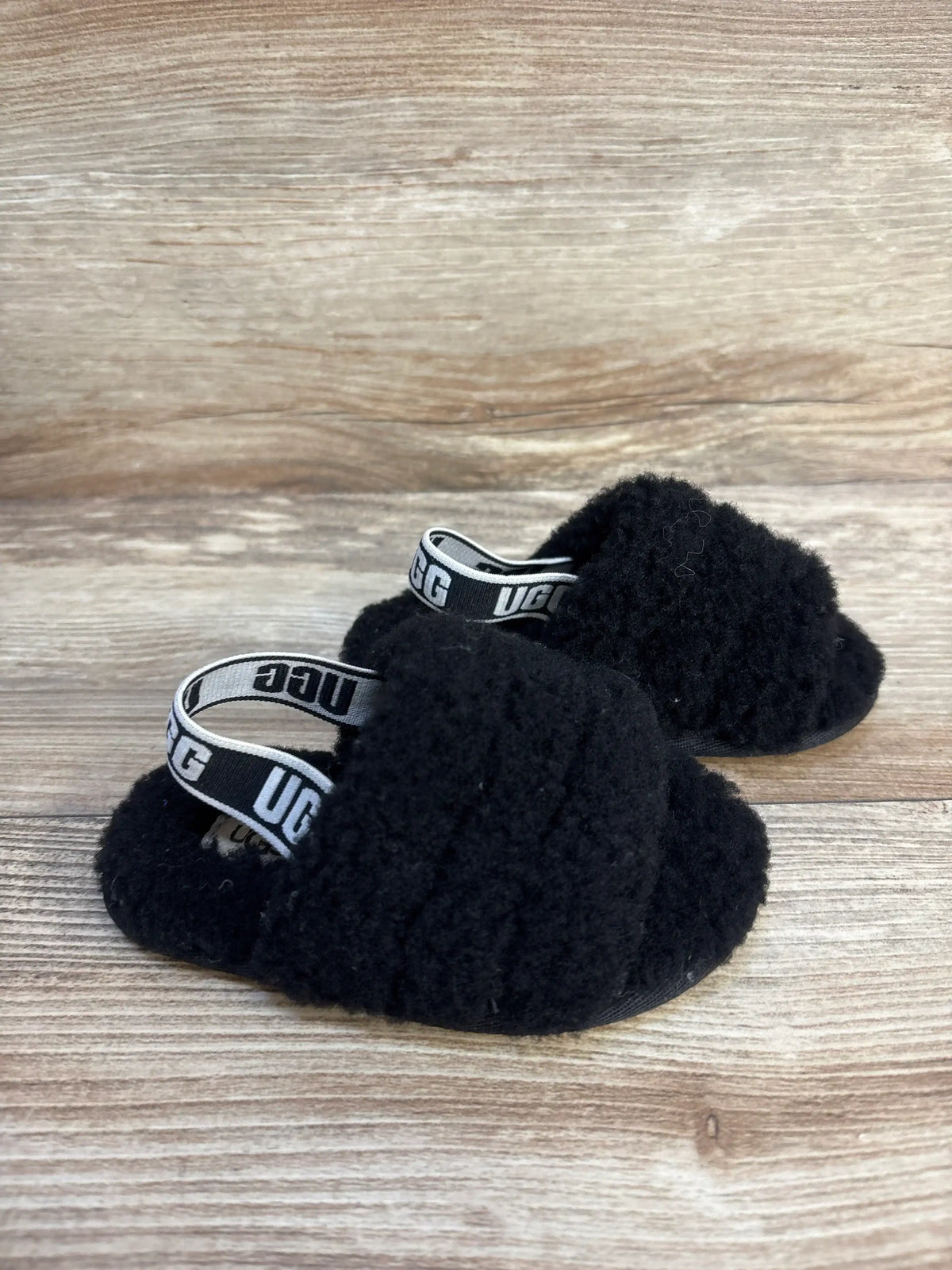 Angled view of UGG Fluff Yeah toddler slides in black showing fuzzy upper and elastic UGG heel strap on wood background