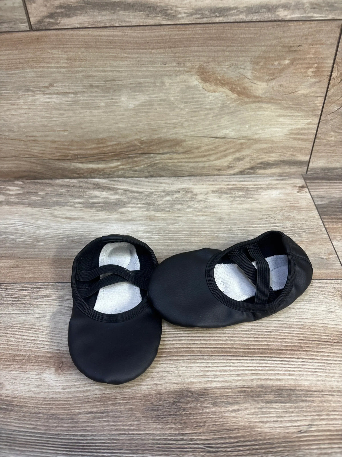 Top-down view of Stelle toddler black ballet shoes size 6/7C showcasing elastic cross straps and white insoles on wood floor