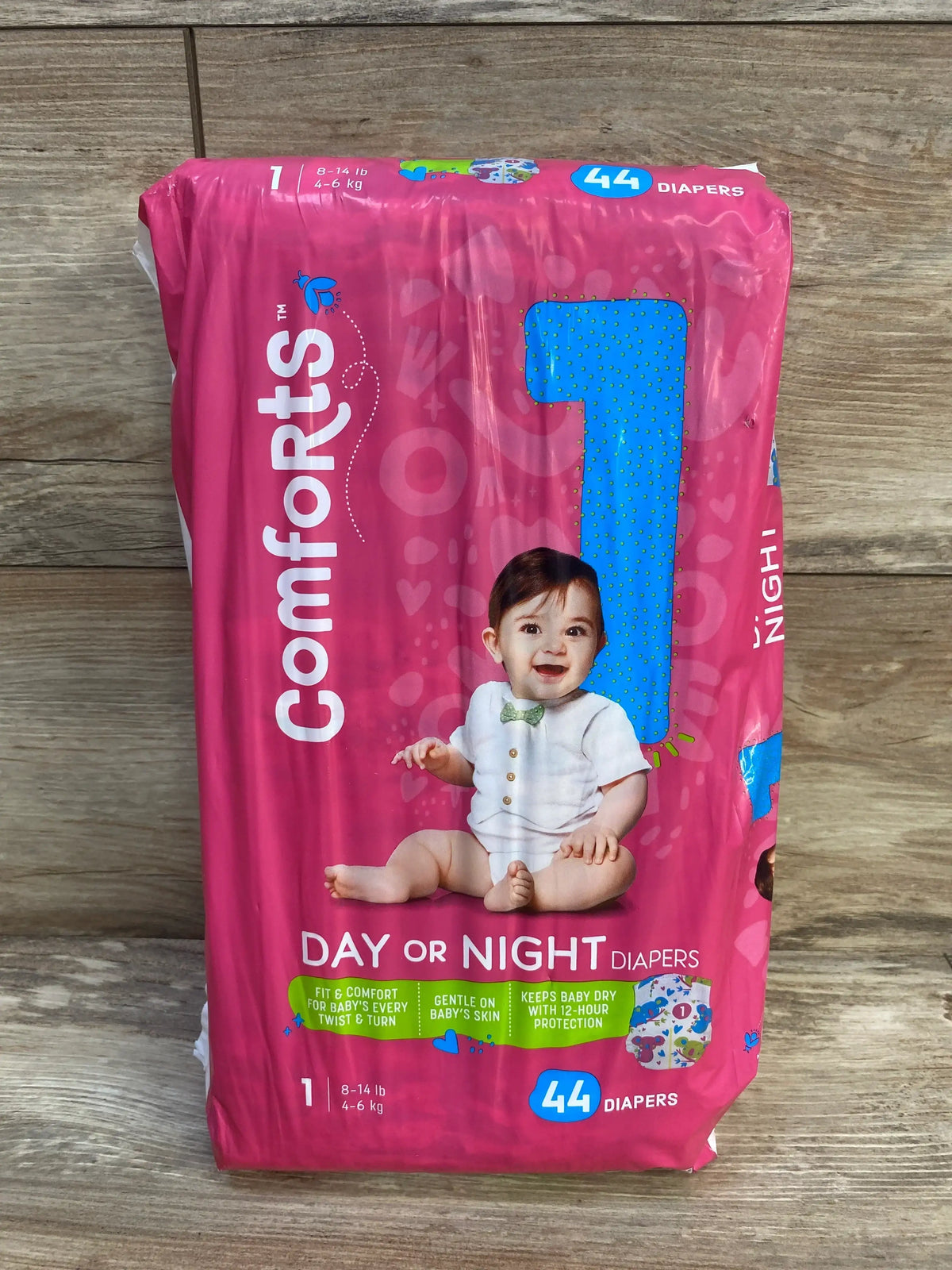 Front view of Comforts size 1 diaper pack showing baby photo, Day or Night text, 44 count and 8 to 14 lb on wood background