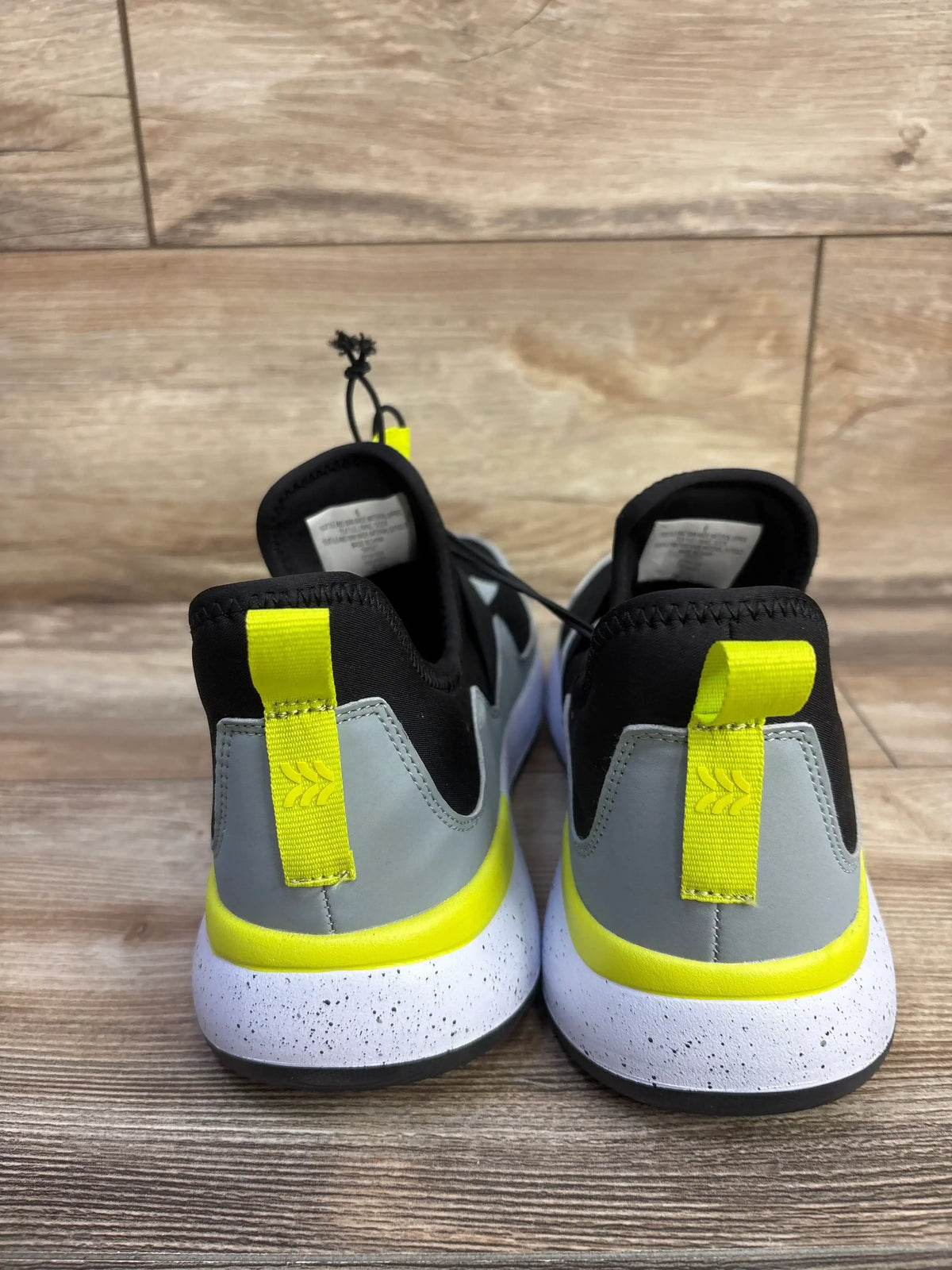 Rear view of All in Motion kids slip on sneakers with neon yellow heel tabs, grey upper and speckled white midsoles on wood floor
