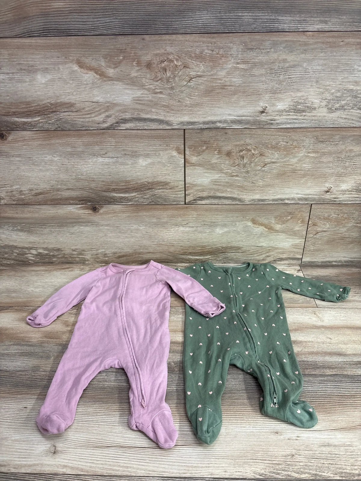 Flat lay of Cloud Island 2-pack sleeper set showing mauve and green footed sleepers, one with pink heart pattern, on wood background