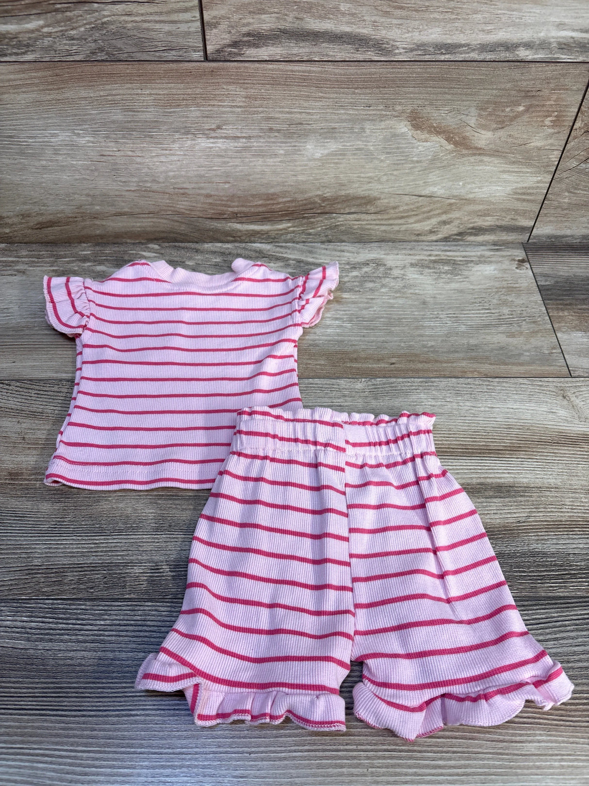 Flat lay of newborn pink ribbed 2-piece shirt and shorts set showcasing red stripes and ruffled sleeves and hems on wood background
