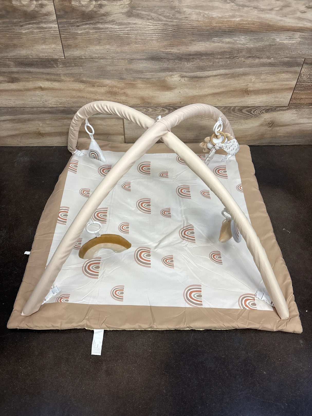 Top-down view of Mallify Tummy Time Play Gym Rainbow showing crossed beige arches, three hanging toys, and rainbow-print padded mat on dark floor