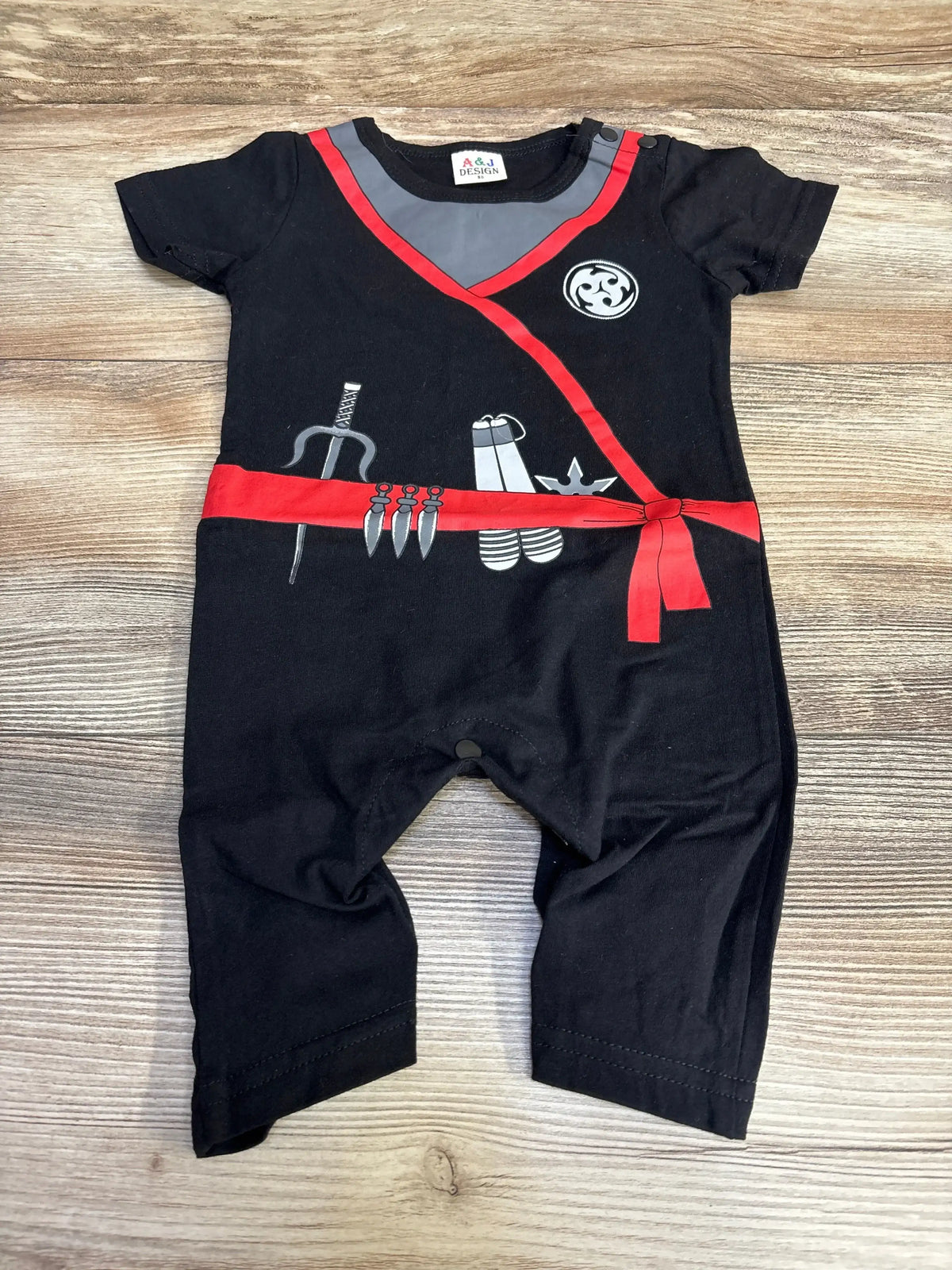 Front view of black ninja romper showcasing red trim, belt, and weapon graphics on a wood background