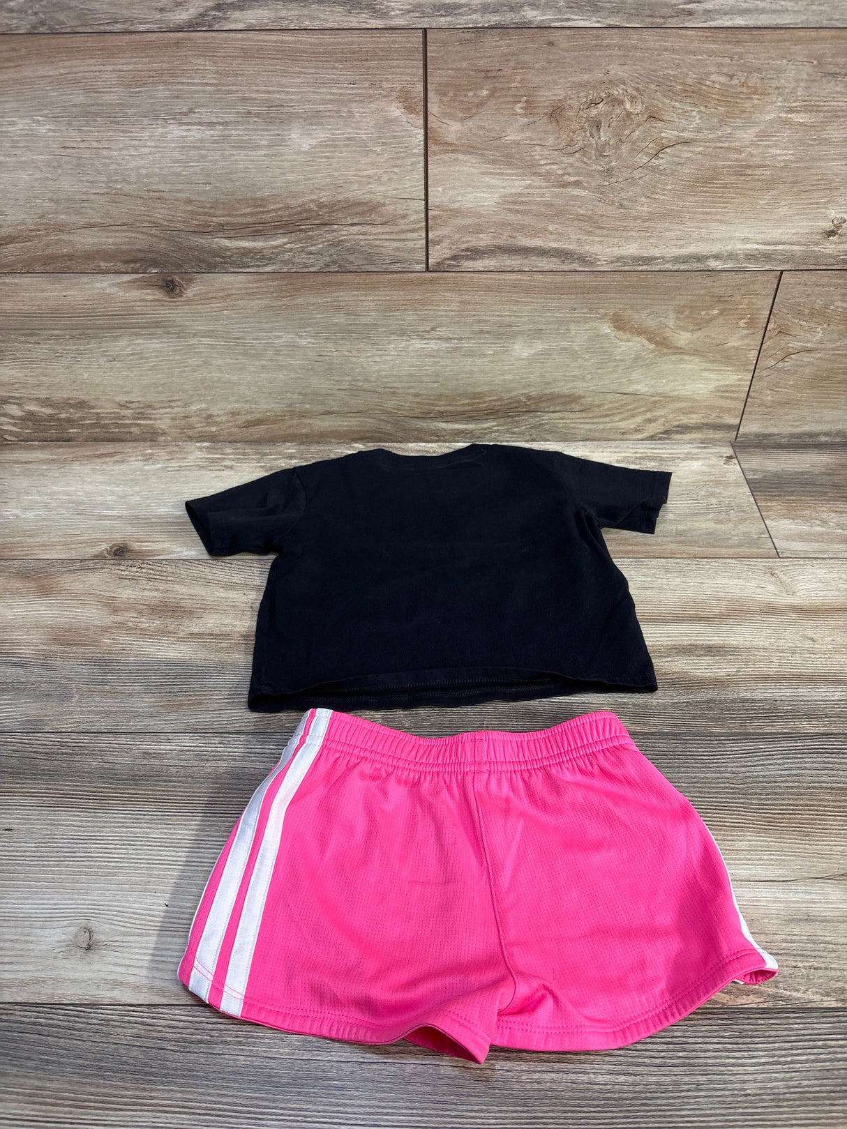Flat lay of Adidas toddler 2-piece set: black cropped T-shirt (back) and pink shorts with white side stripes on wood floor