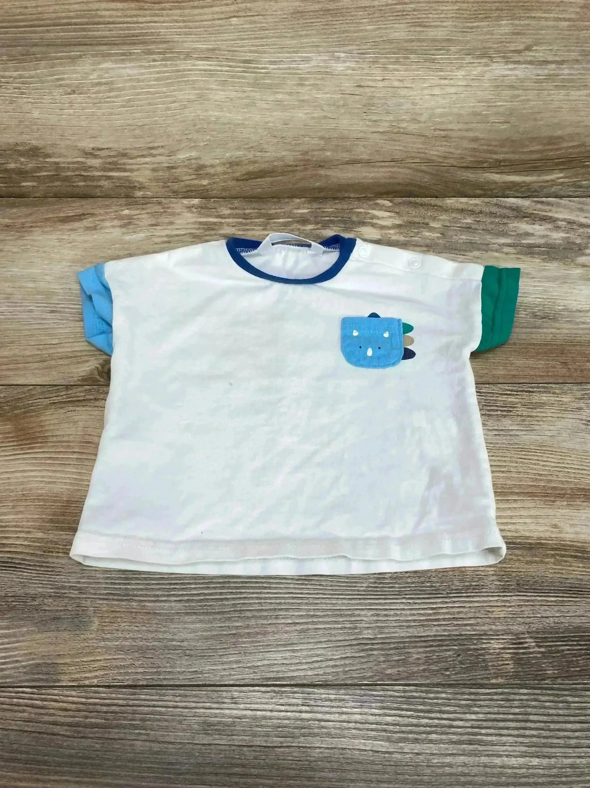 Flat lay of Mayoral Dino Pocket Shirt size 2-4m showcasing white body with navy trim blue and green sleeves and dinosaur chest pocket on wood plank floor