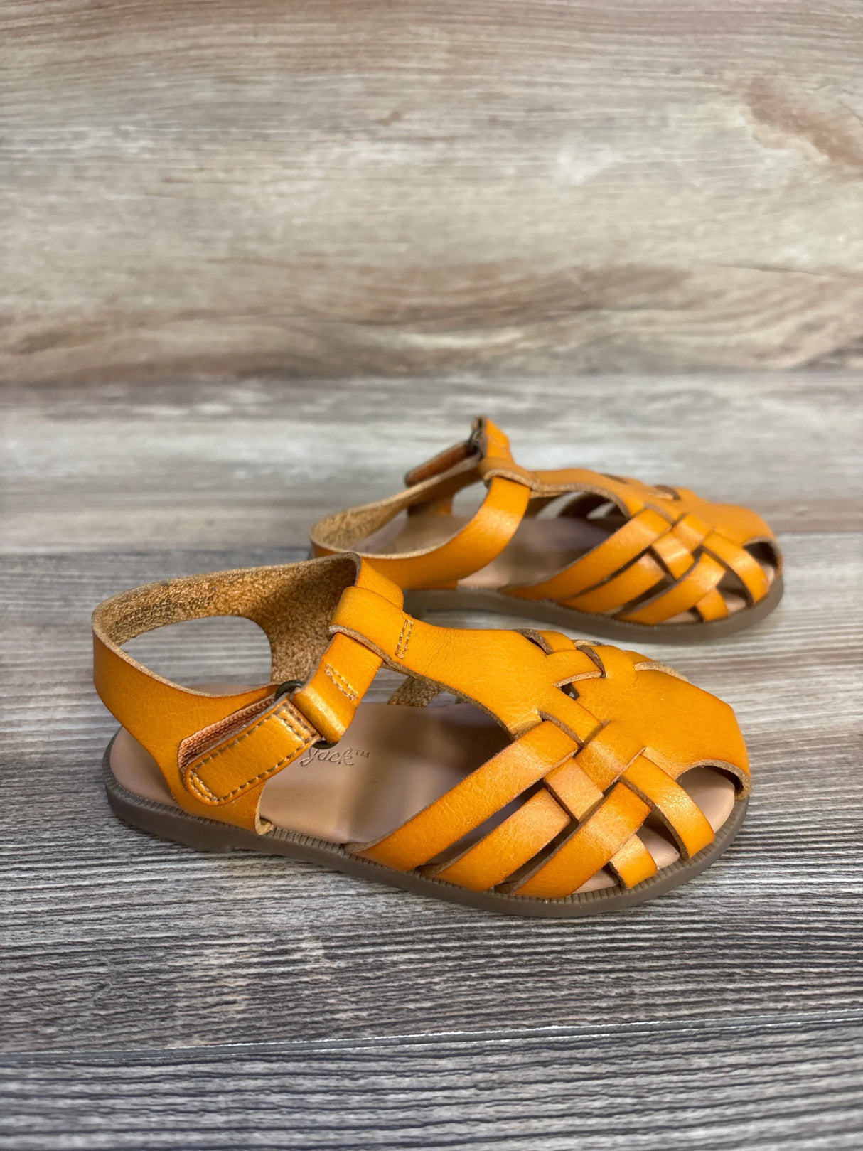 Three-quarter view of Cat & Jack Ella Ankle Strap Sandal in cognac sz 7c showcasing woven fisherman straps and ankle strap with wood plank background