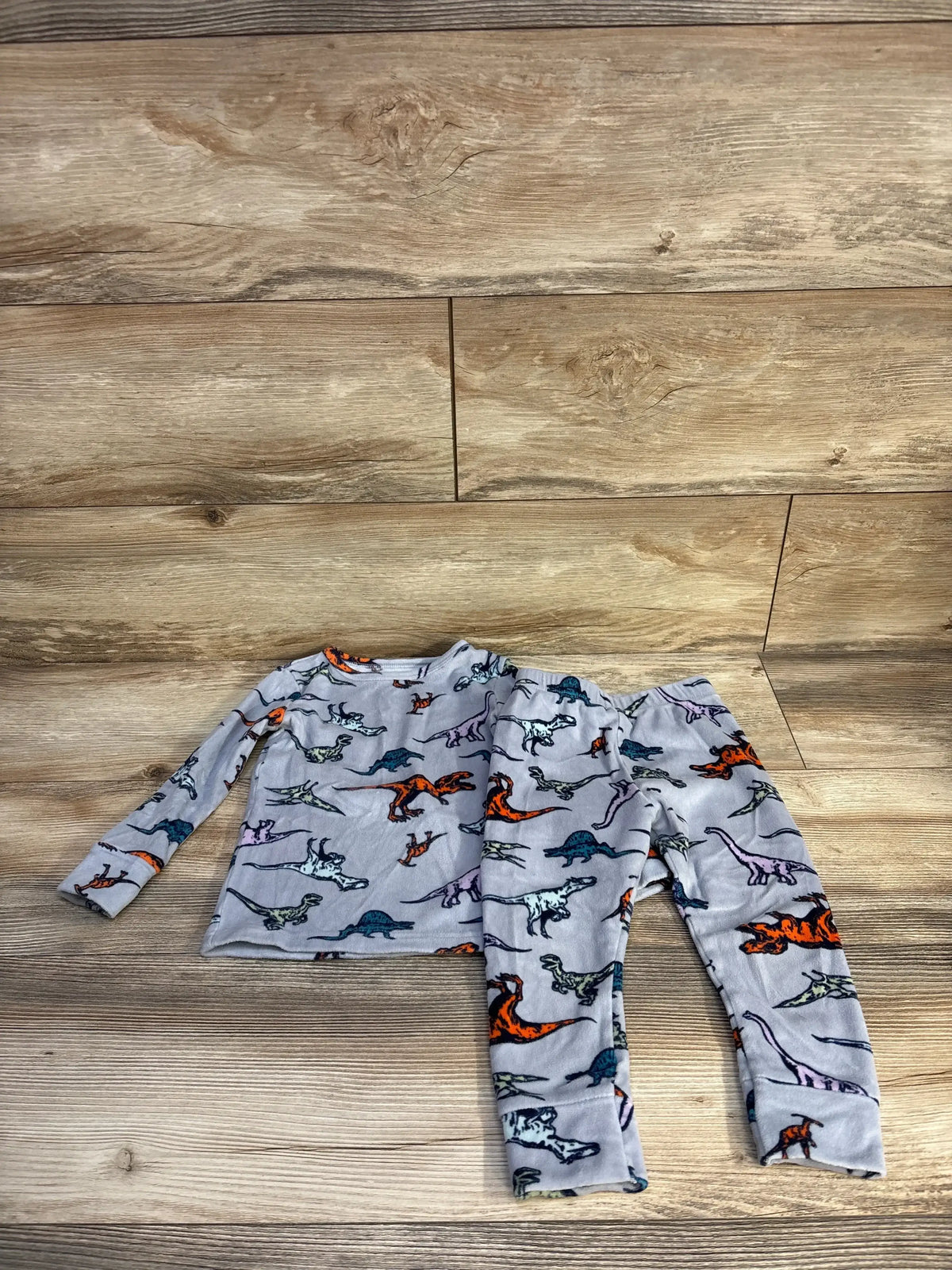Flat lay of Cat and Jack 2pc Dinosaur Velour Pajama Set Grey sz 2T showcasing long-sleeve top and matching pants with colorful dinosaur print on wood plank background