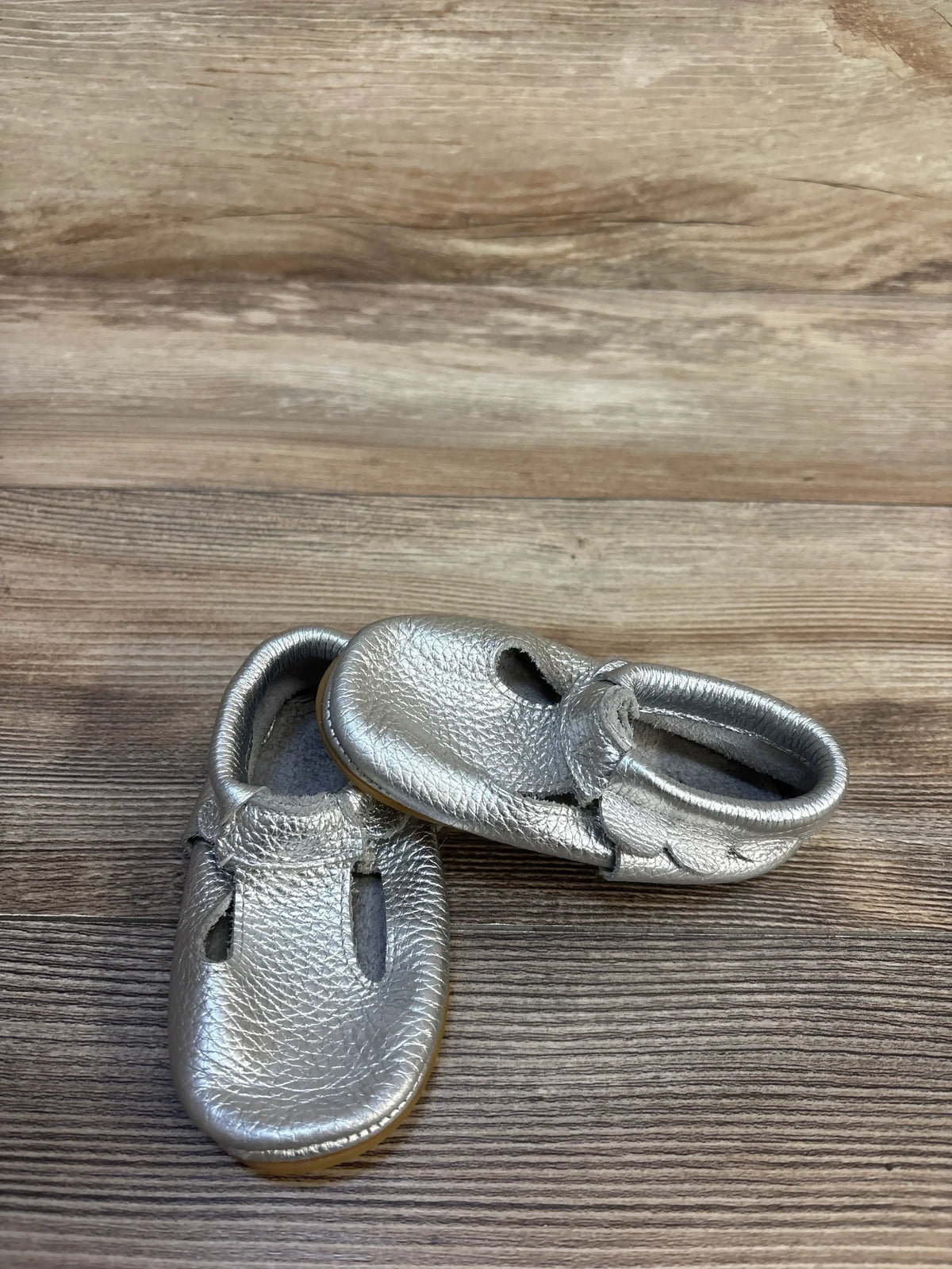 Angled top view of silver leather moccasin baby shoes size 5C showcasing pebbled metallic finish, T-strap cutouts and elasticized heel on light wood background