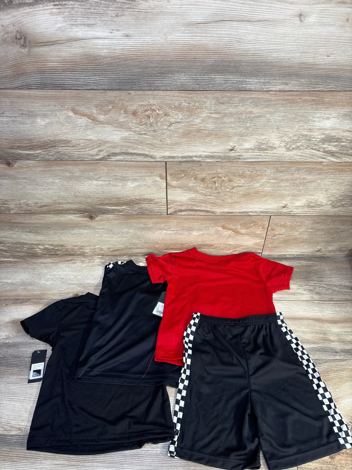 Flat lay of toddler shirt and short set: red and black tees with black shorts with checkerboard side stripes on wood floor
