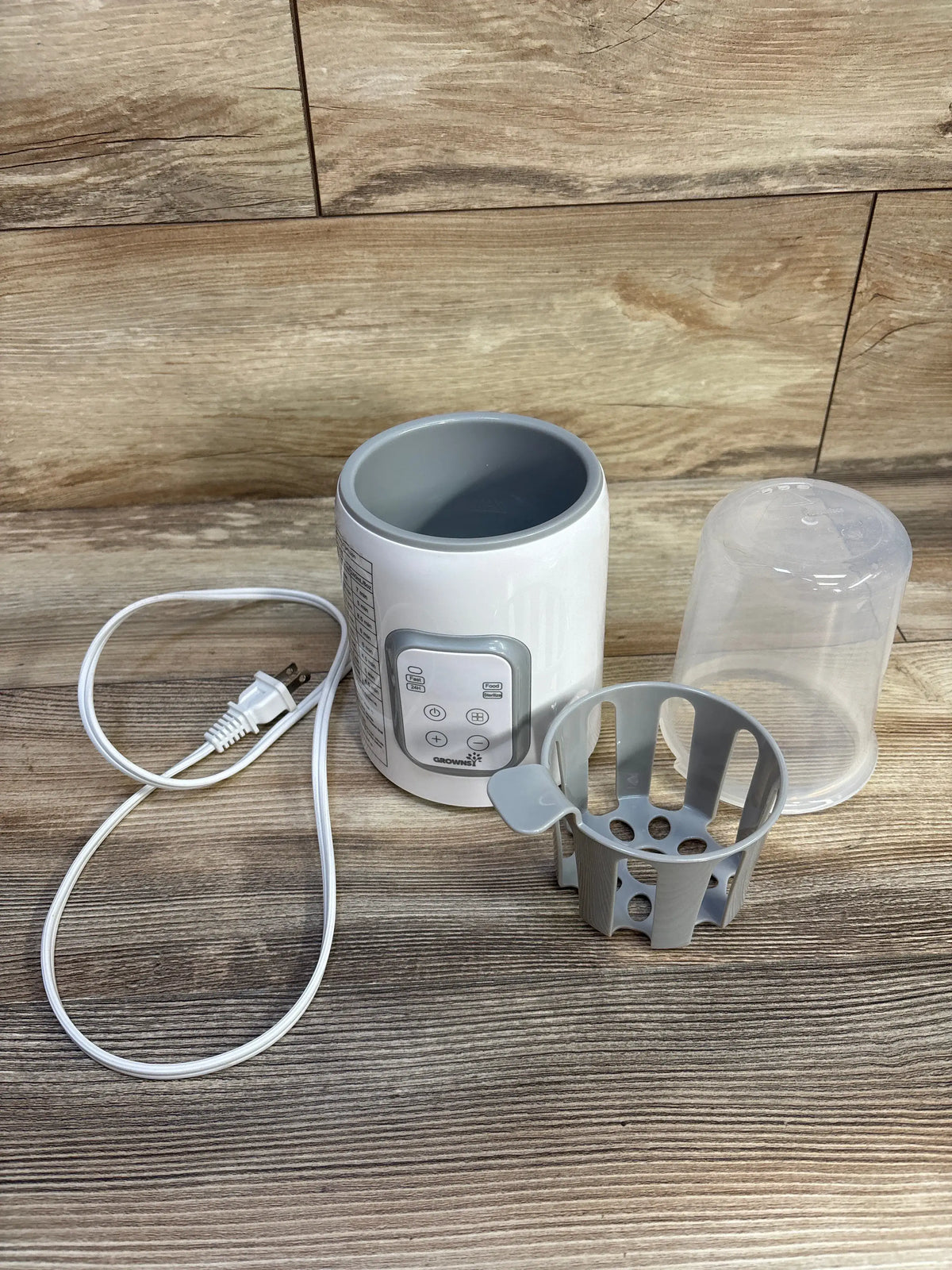 Front view of Grownsy white baby bottle warmer with anti-scald basket, clear cover, and power cord on wood-grain surface