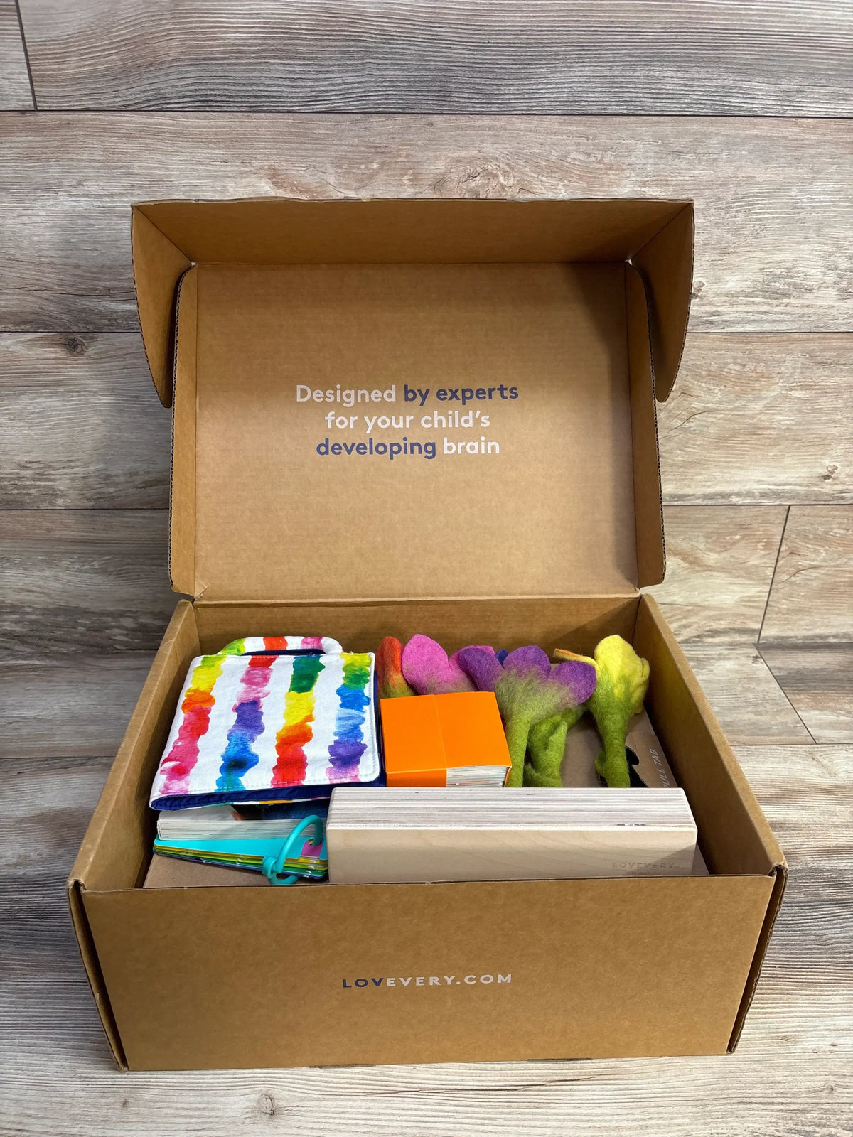 Top view Lovevery Helper Play Kit with colorful toys; lid reads 'Designed by experts for your child's developing brain'