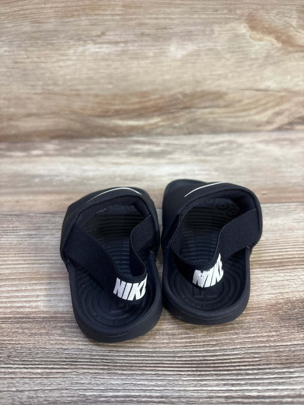 Top-down view of toddler Nike Kawa slides showing white Nike heel logos and contoured ribbed footbeds on wood floor