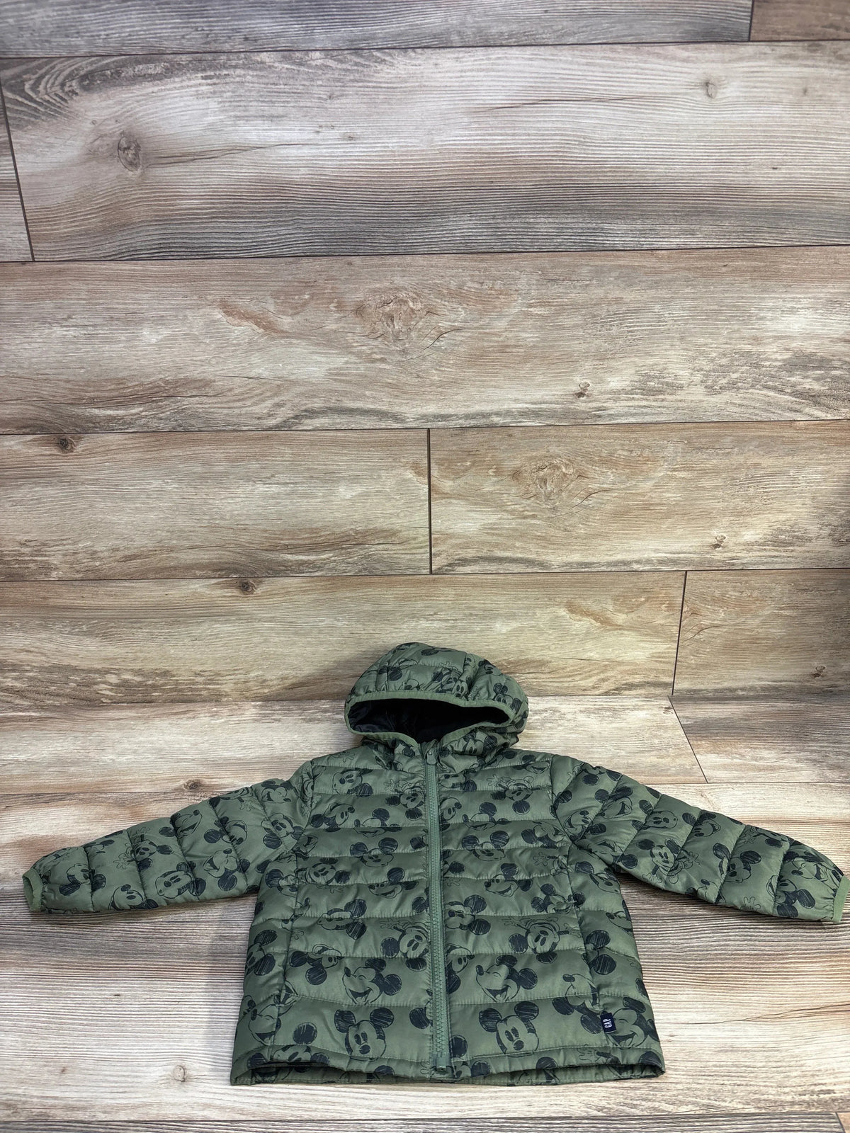 Front view of green puffer jacket with hood, front zipper, and all-over Mickey Mouse print on wood background