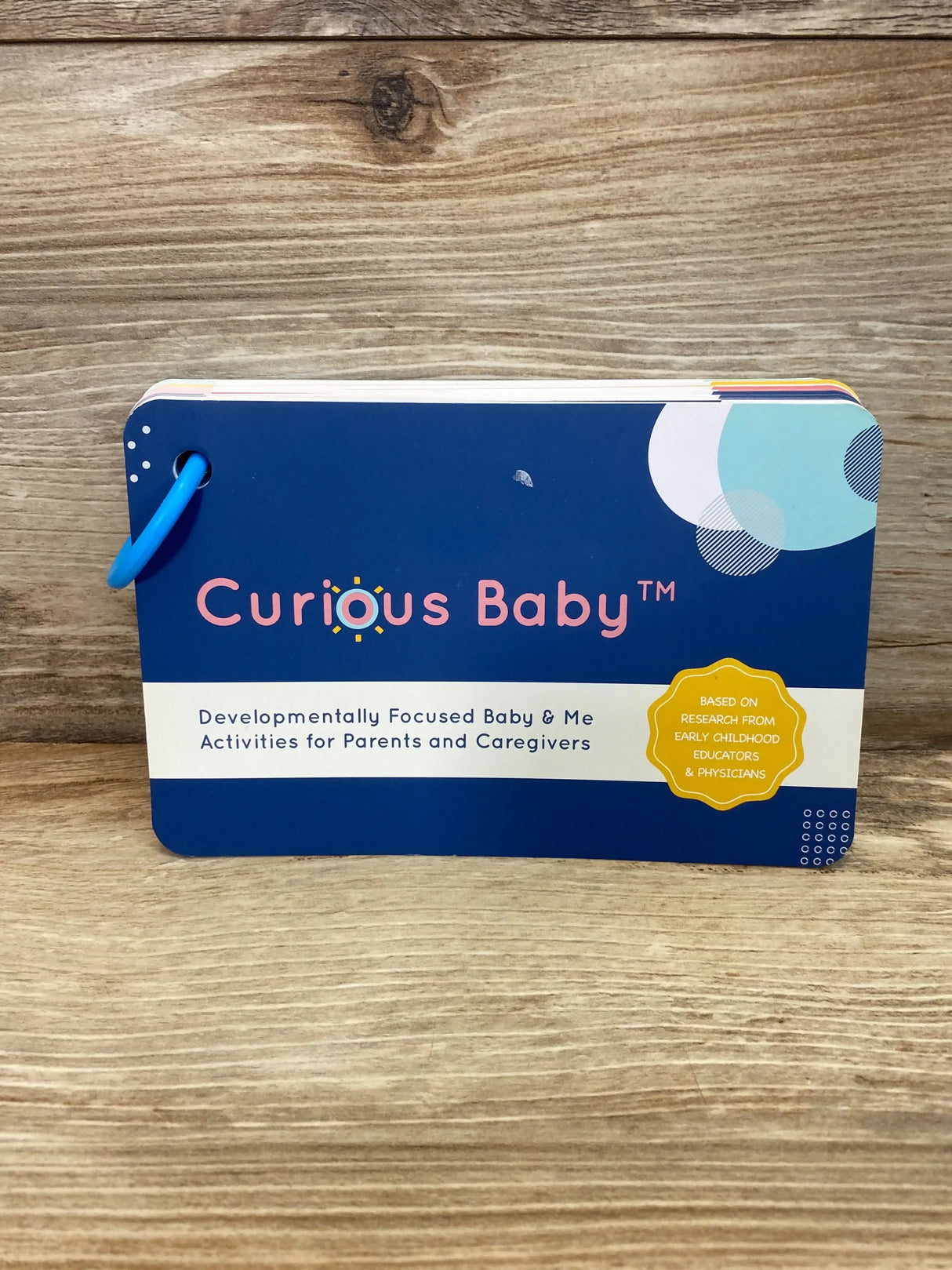 Front view of Curious Baby activity cards with navy cover, white subtitle band and yellow research badge on wooden surface