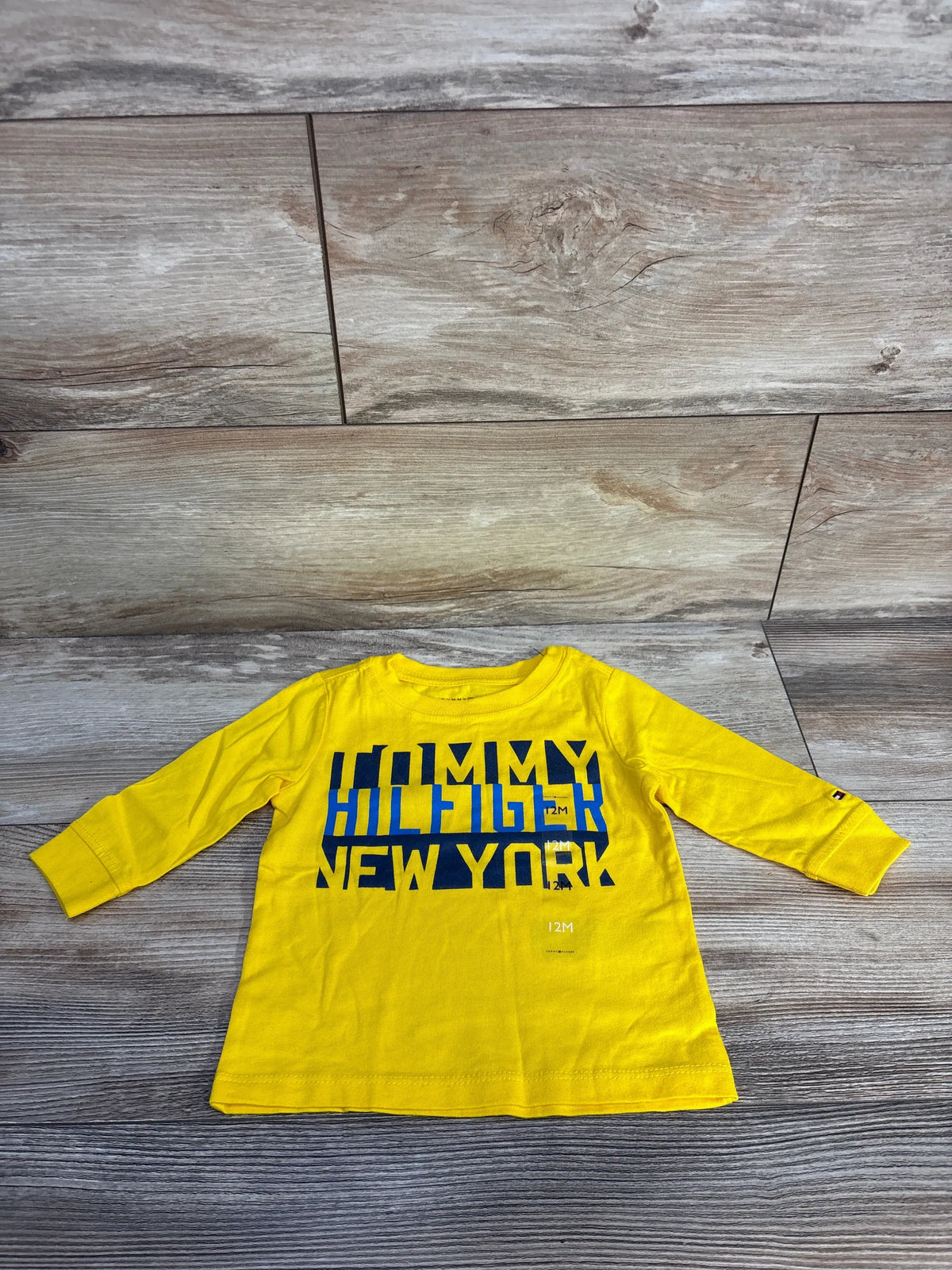 Front view of bright yellow Tommy Hilfiger logo shirt size 12 months with long sleeves and embroidered chest logo on a wood floor