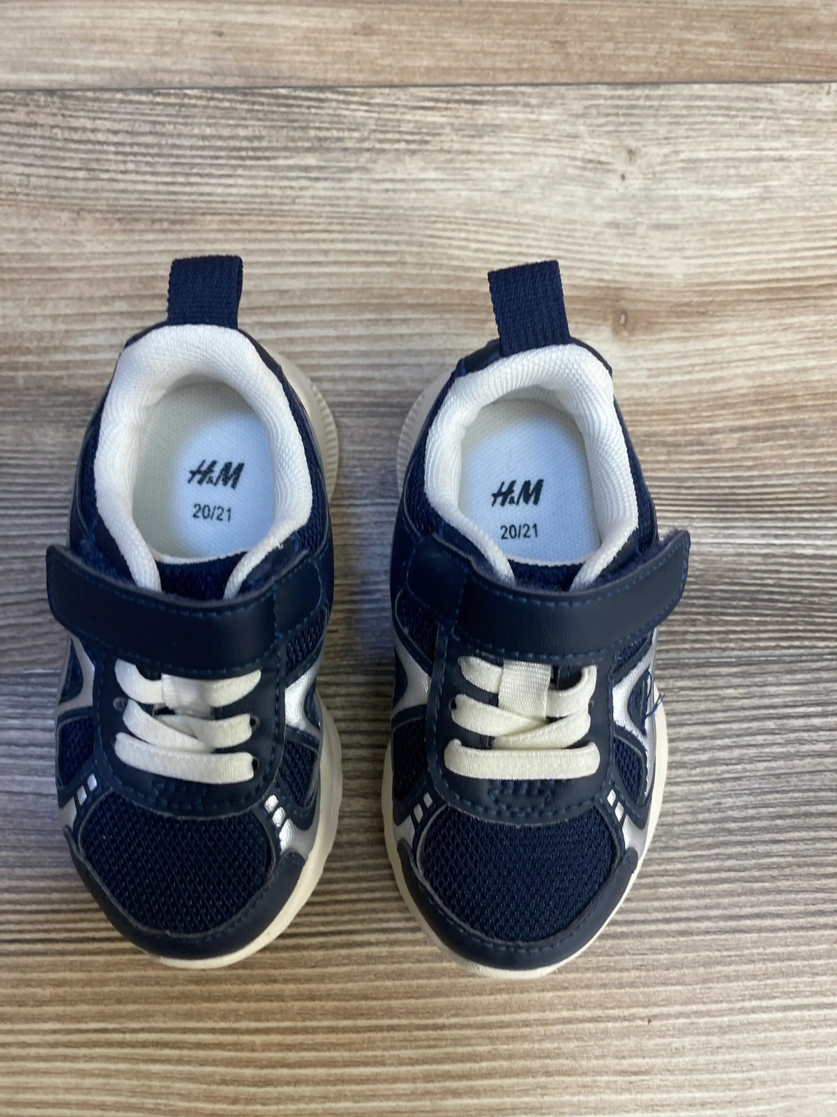Top-down view of H&M navy toddler sneakers 20/21 with velcro strap, elastic laces, mesh upper and white sole on wood background