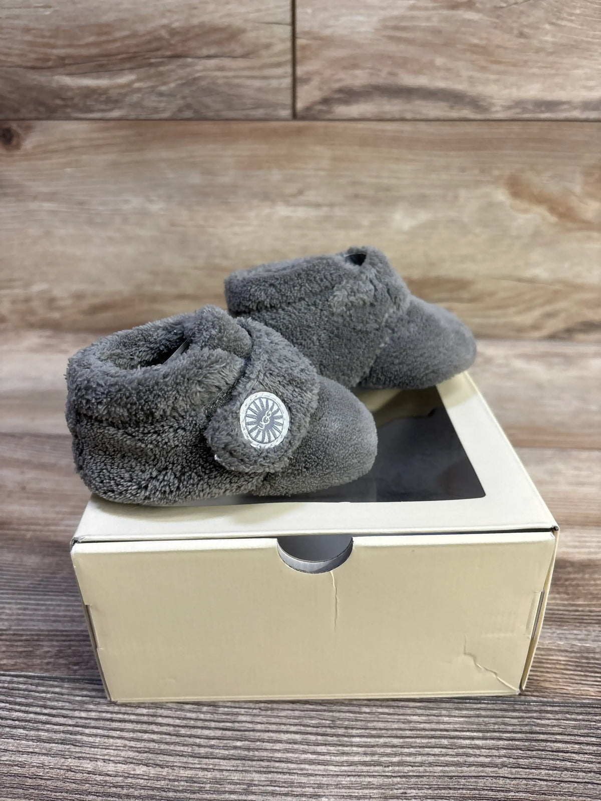 Three-quarter view of UGG Bixbee baby booties in grey showing plush fleece exterior and strap with logo on a shoebox and wood backdrop