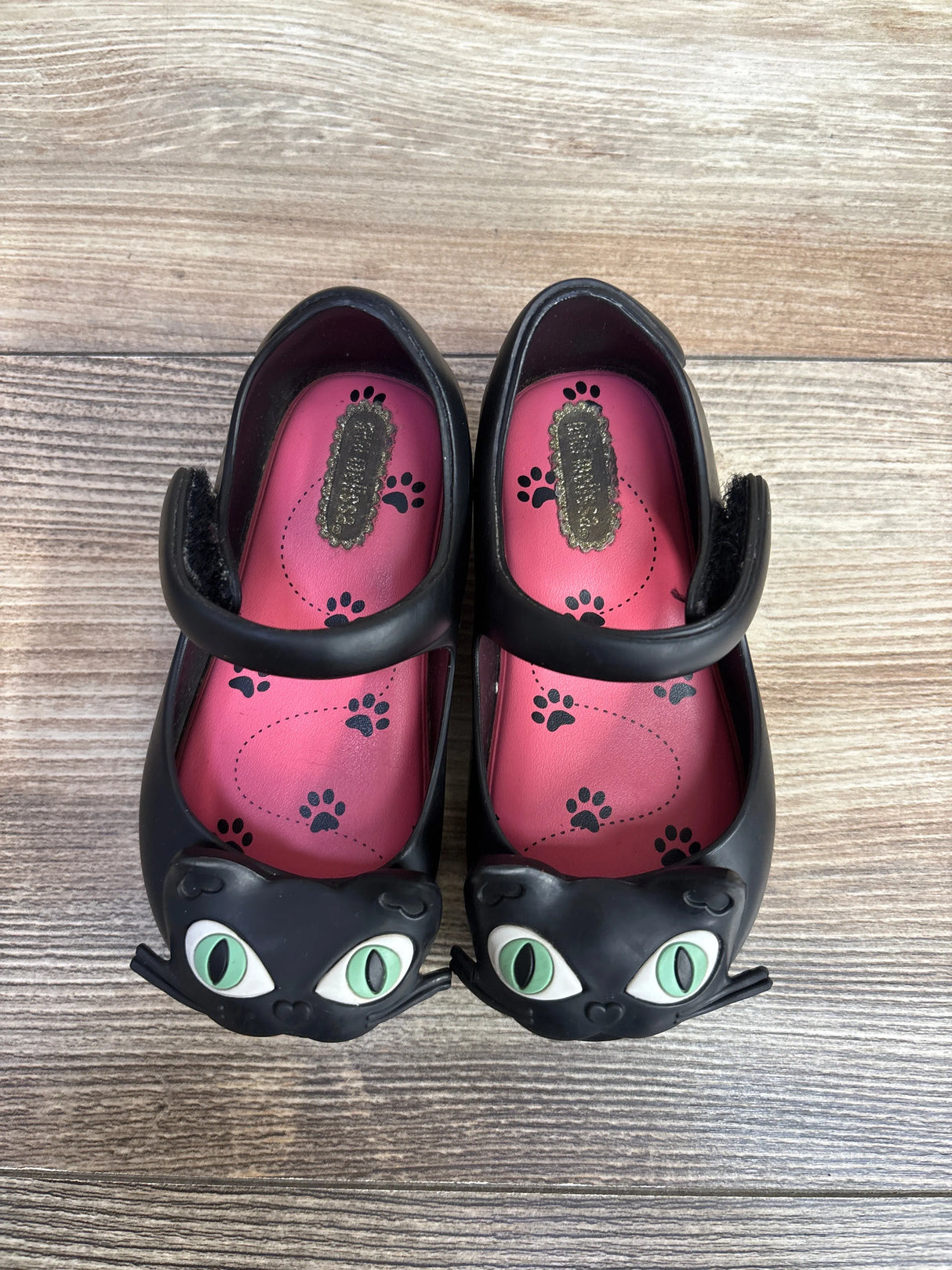 Pair of black girls' Mary Jane shoes with green cat eyes and pink paw-print insoles on wood floor