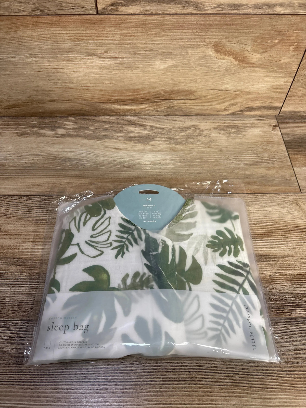Front view of Little Unicorn sleep sack (6-12 months) with green tropical leaf print in clear packaging on wood background