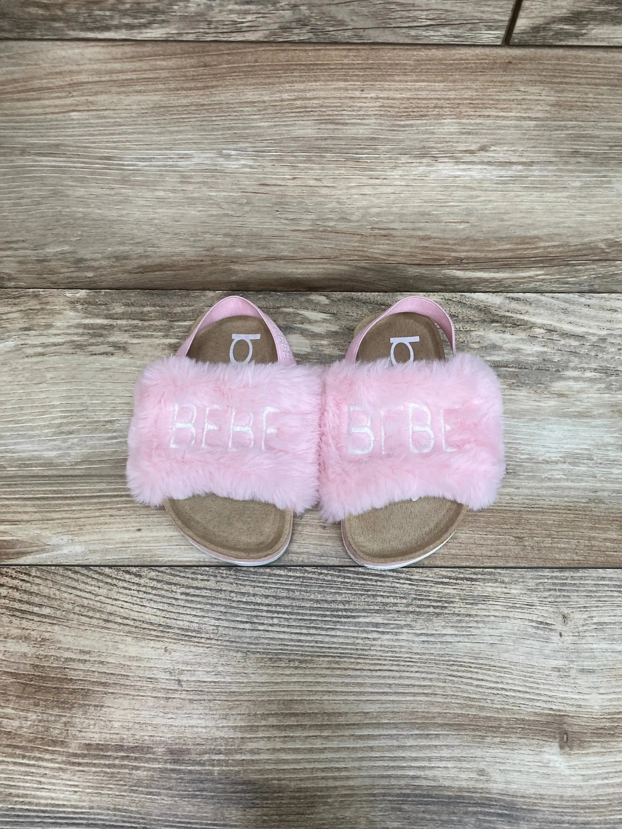 Top-down view of pink faux-fur Bebe slide sandals showing embroidered BEBE on the straps on a wood floor background