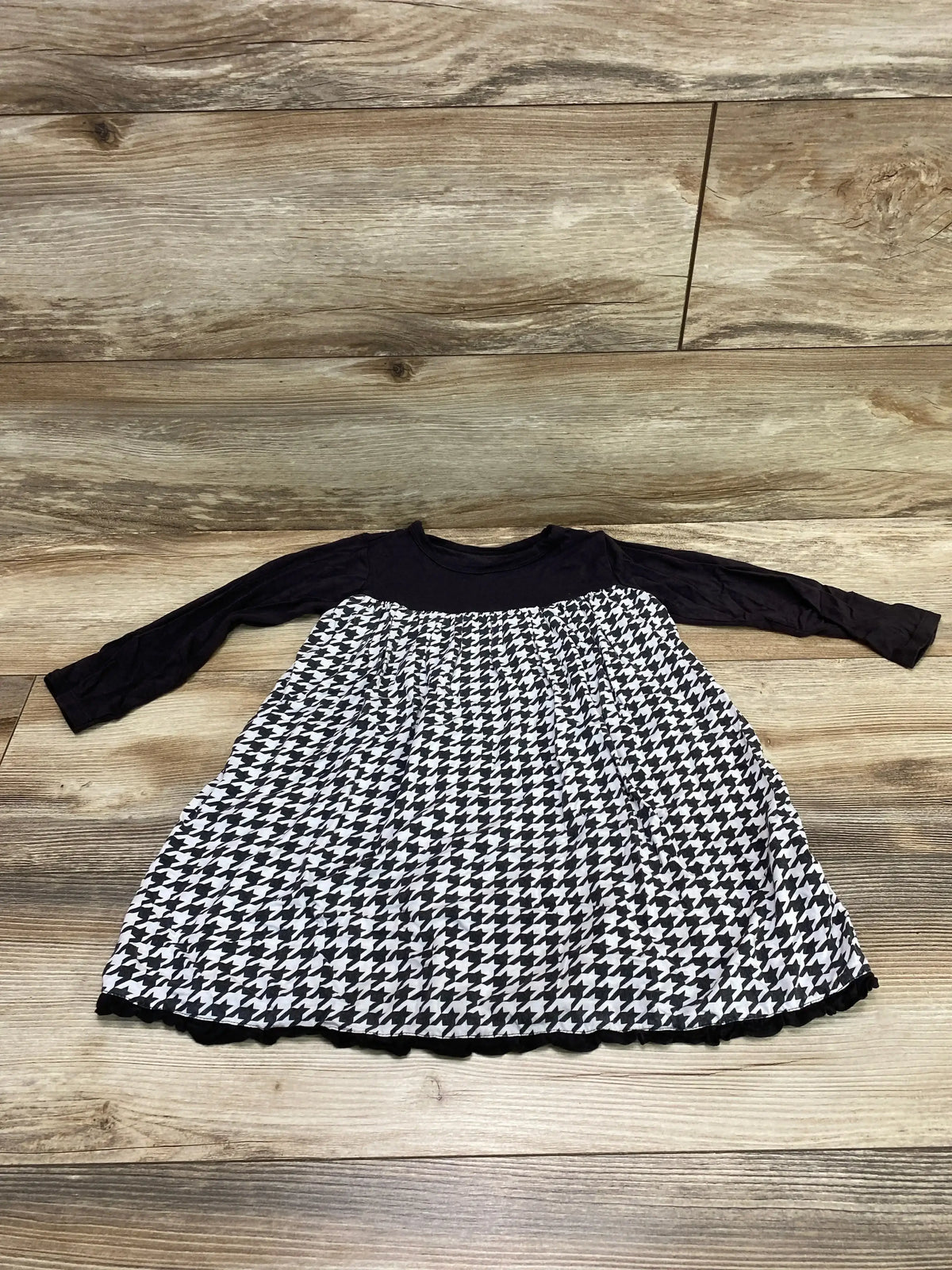 Front flat lay of Kickee Pants toddler dress showcasing black long-sleeve bodice and gathered black-and-white houndstooth skirt on wood floor