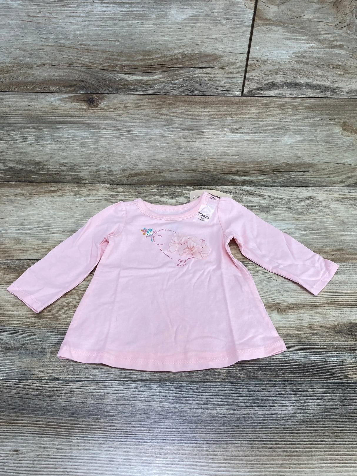 Front flat-lay of pink long-sleeve baby shirt (3-6 months) showing birdie embroidery with 3D flower on wood background