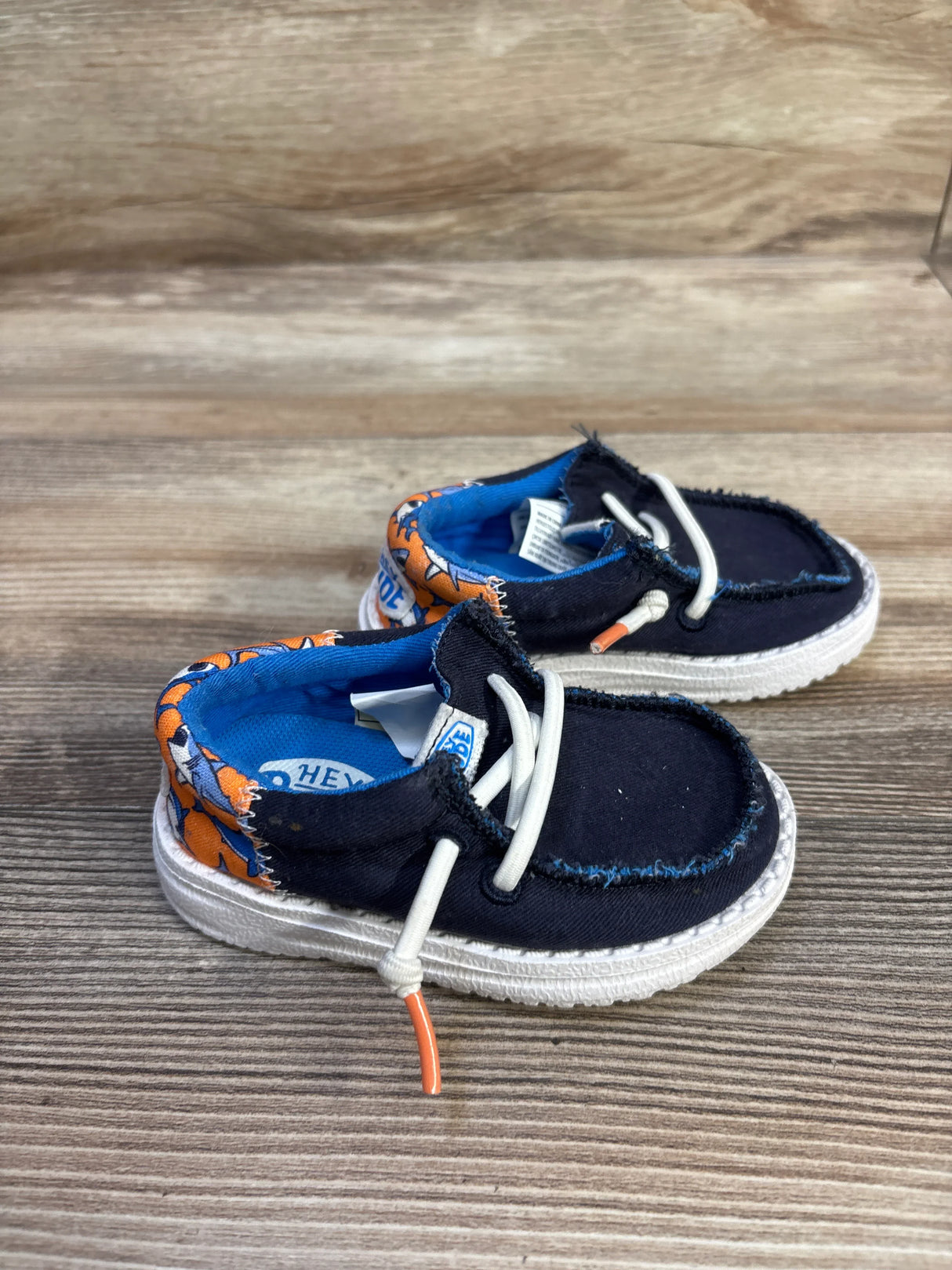 Three-quarter view of Hey Dude Wally navy shoes with orange shark-print heel lining and white elastic laces on wood