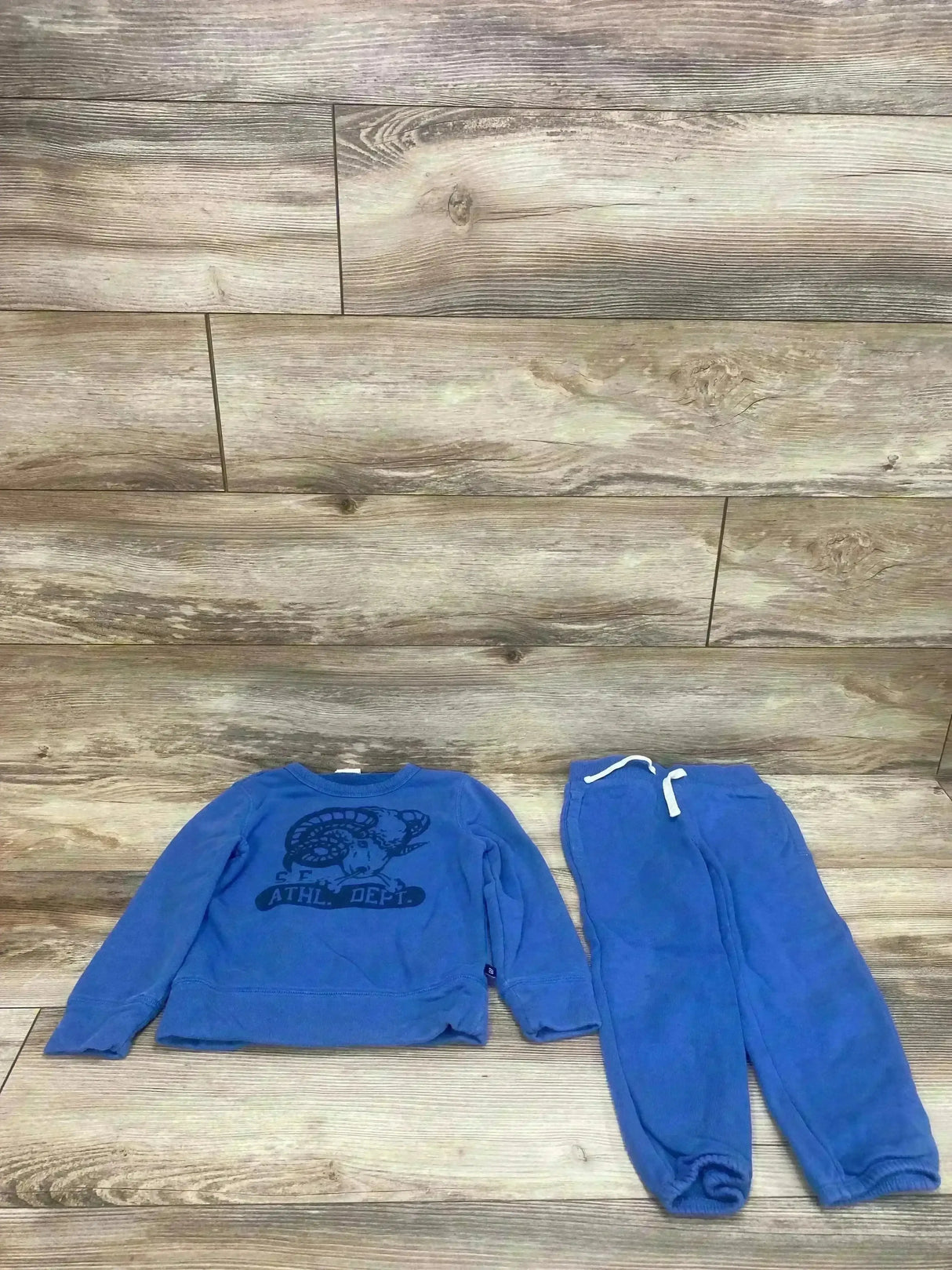 Flat lay of blue Gap Kids sweatshirt and joggers showing crewneck ram graphic and drawstring elastic waist on wood floor