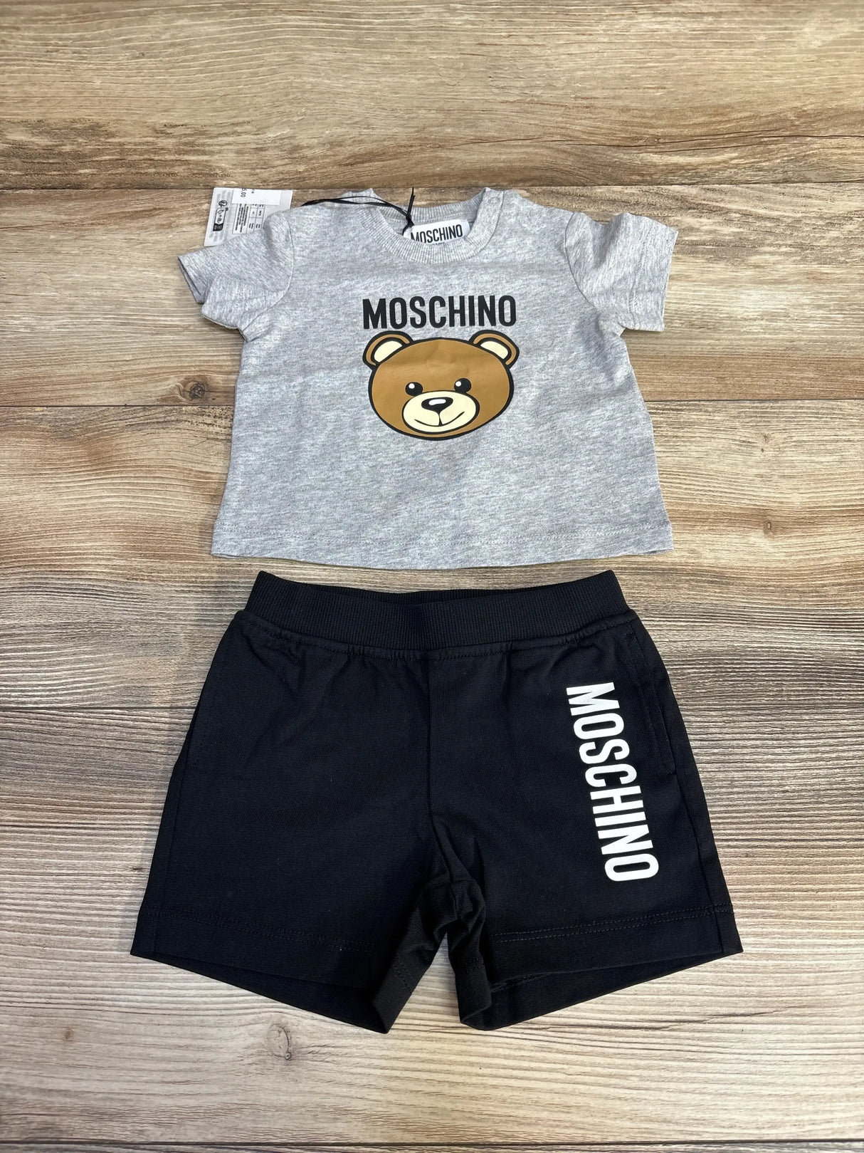 Flat lay of Moschino two-piece set: grey tee with bear print and black shorts with MOSCHINO logo on wood background