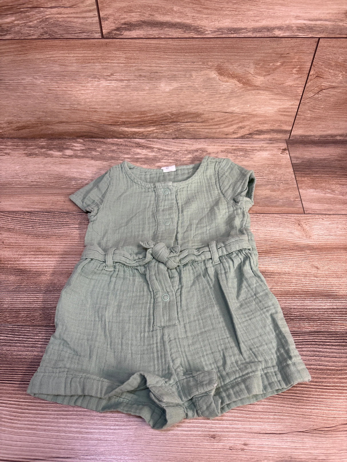 Top-down flat lay of light green Baby Gap muslin shortie romper with front snaps and tied waist belt on wood background