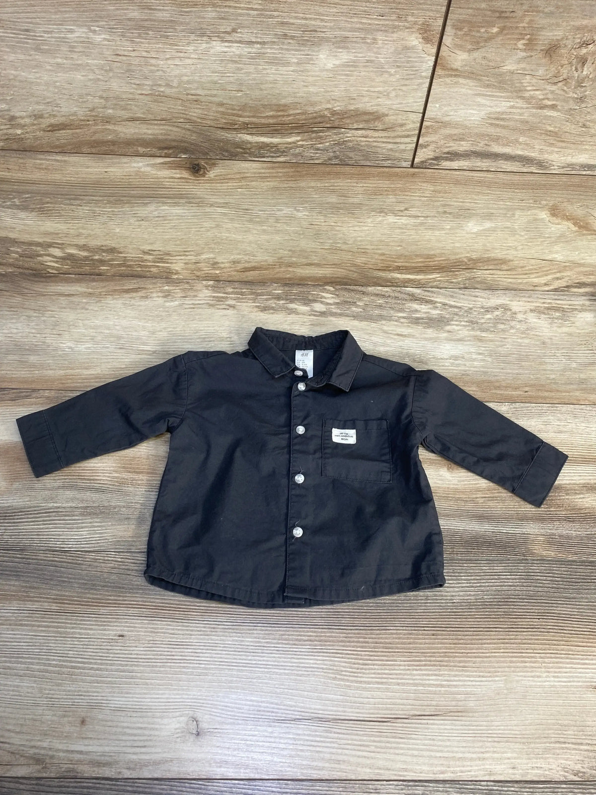 Black long sleeve button-up shirt for toddlers with front pocket, laid flat on wood floor