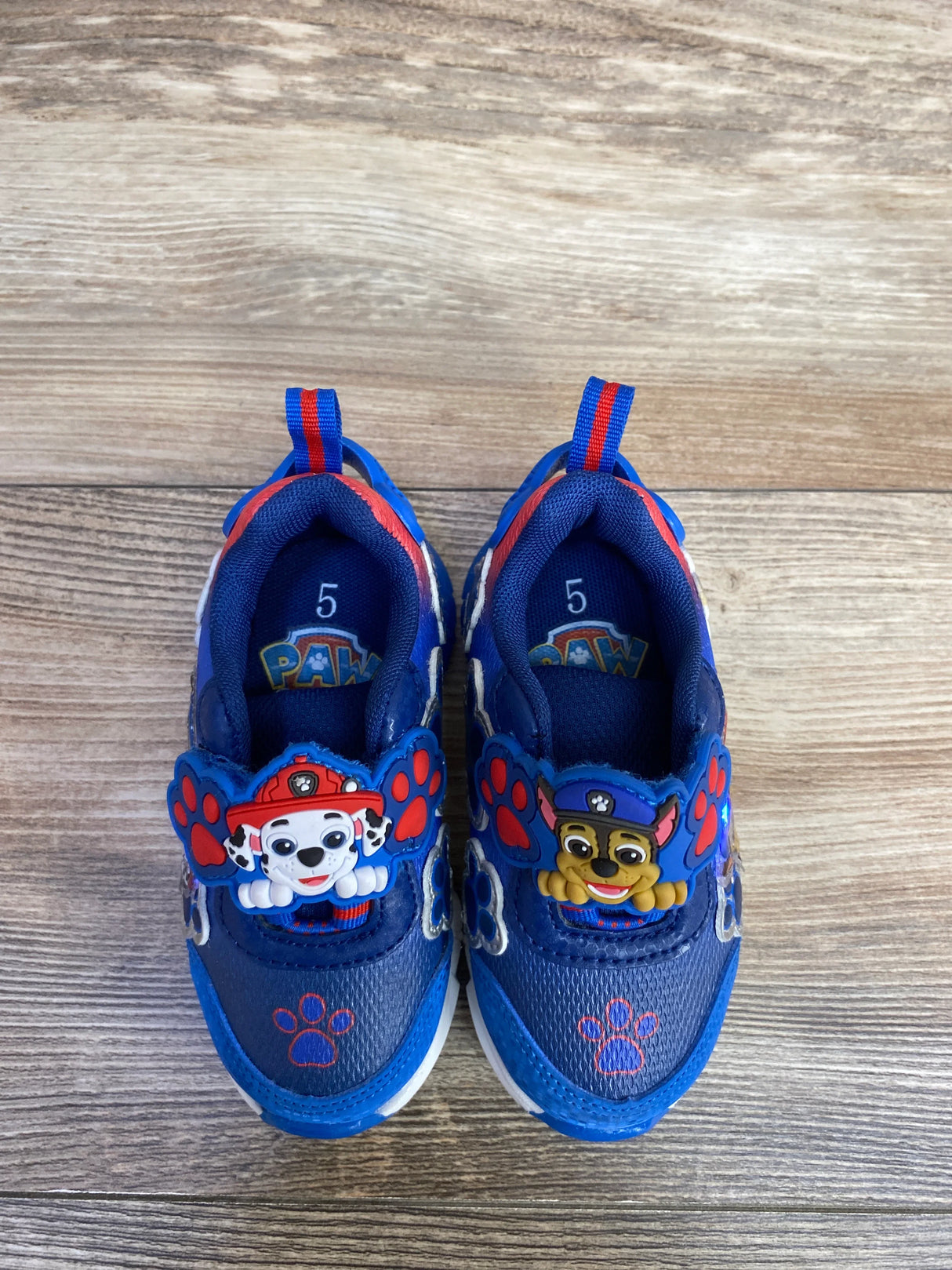 Blue toddler sneakers featuring Paw Patrol characters Marshall and Chase with red paw prints on wooden floor