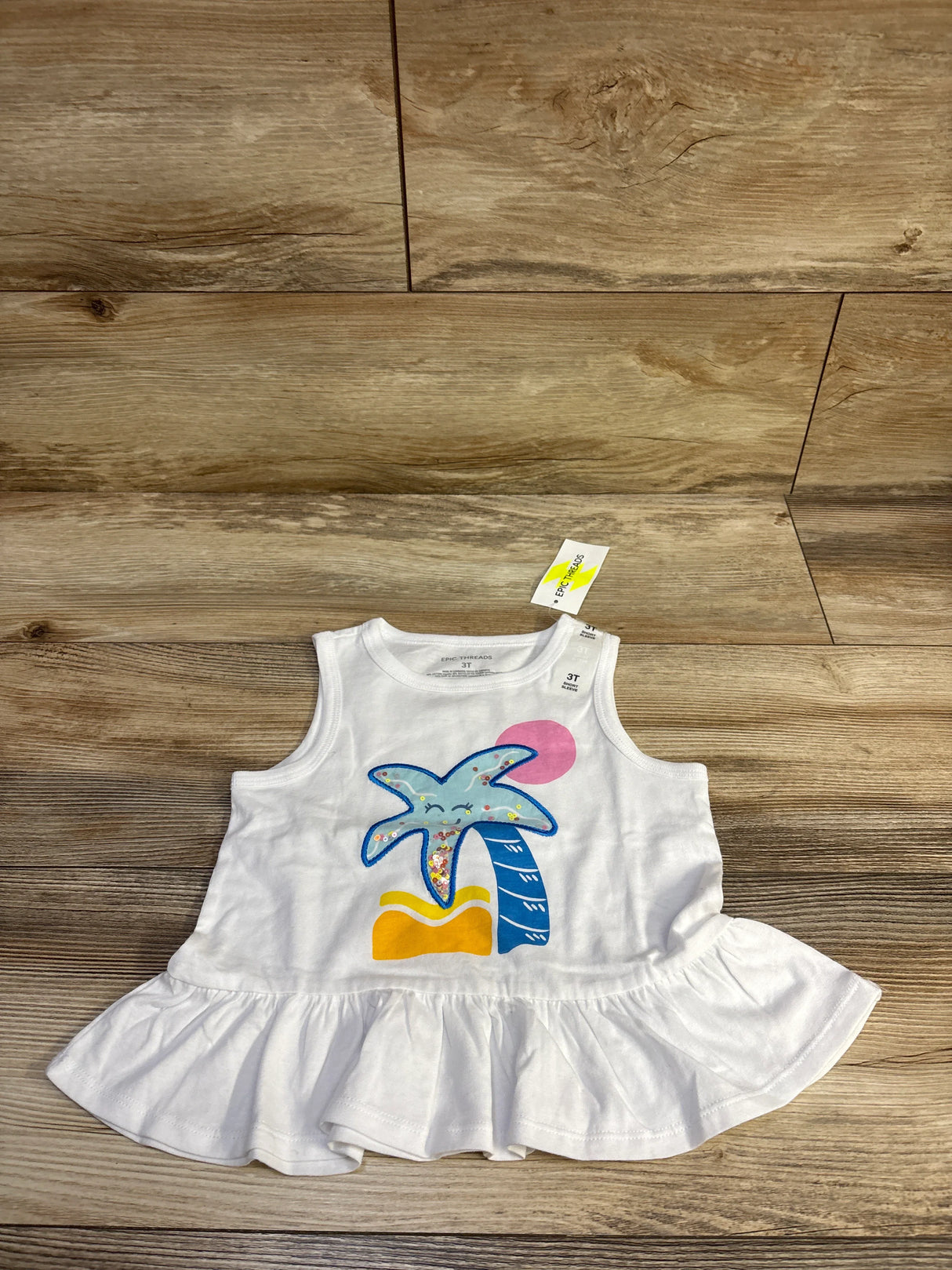 Front flat lay of Epic Threads white peplum tank top showing smiling palm tree graphic and ruffle hem on wood background