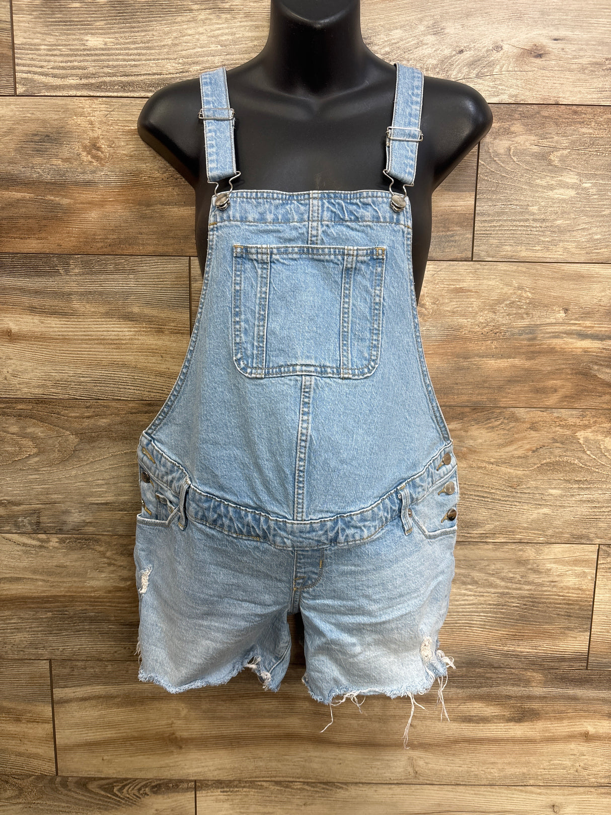 Front view of light blue maternity denim shortalls with straps, bib pocket, side buttons and frayed hem on wood