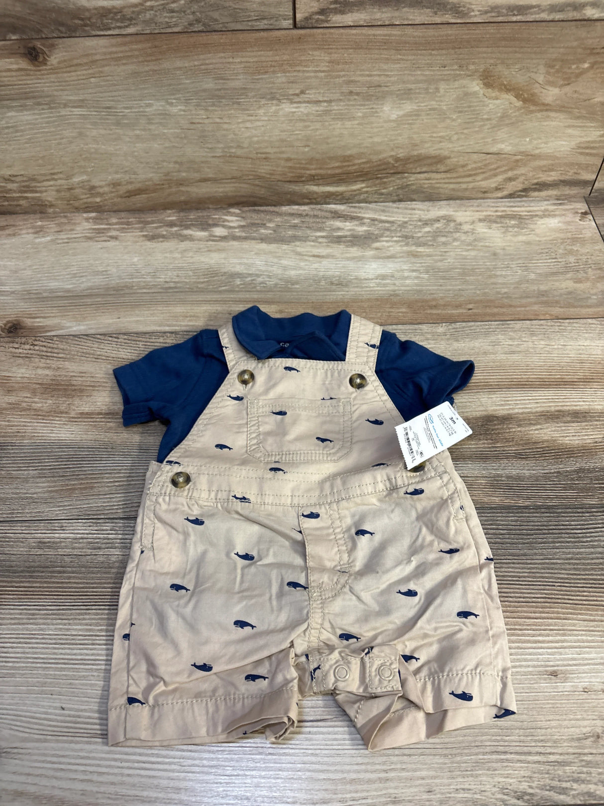 Flat lay of 2-piece navy polo and beige whale-print shortalls with front pocket, buttons and snap crotch on wood background