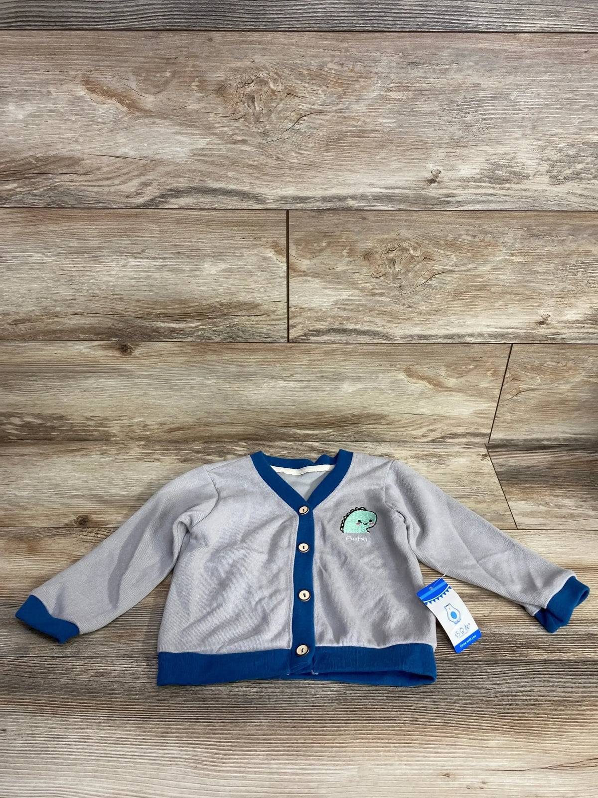 Gray baby cardigan with blue trim and dinosaur patch, laid flat on wood background