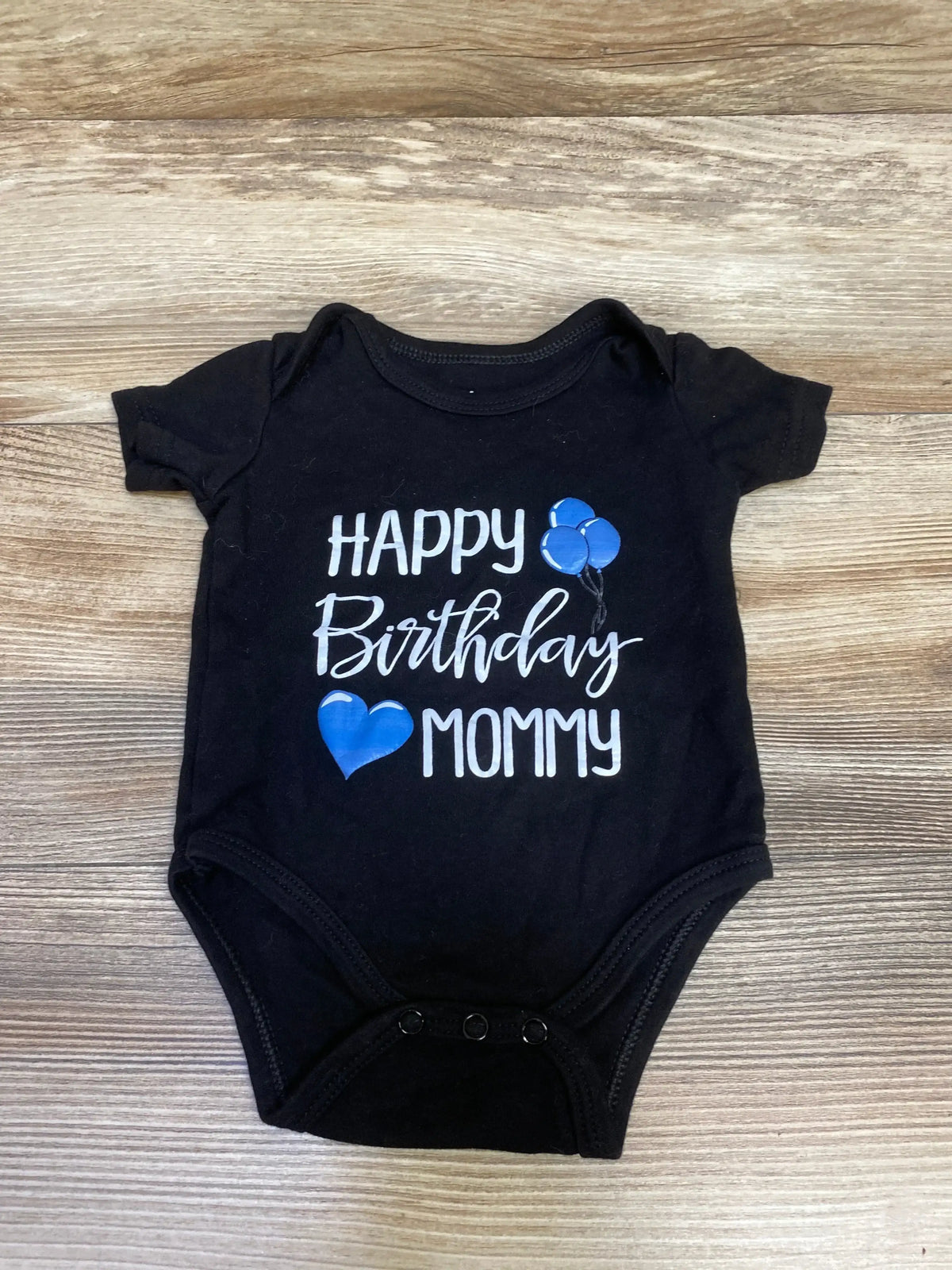 Black baby onesie with white text Happy Birthday Mommy and blue balloons and heart on wooden background