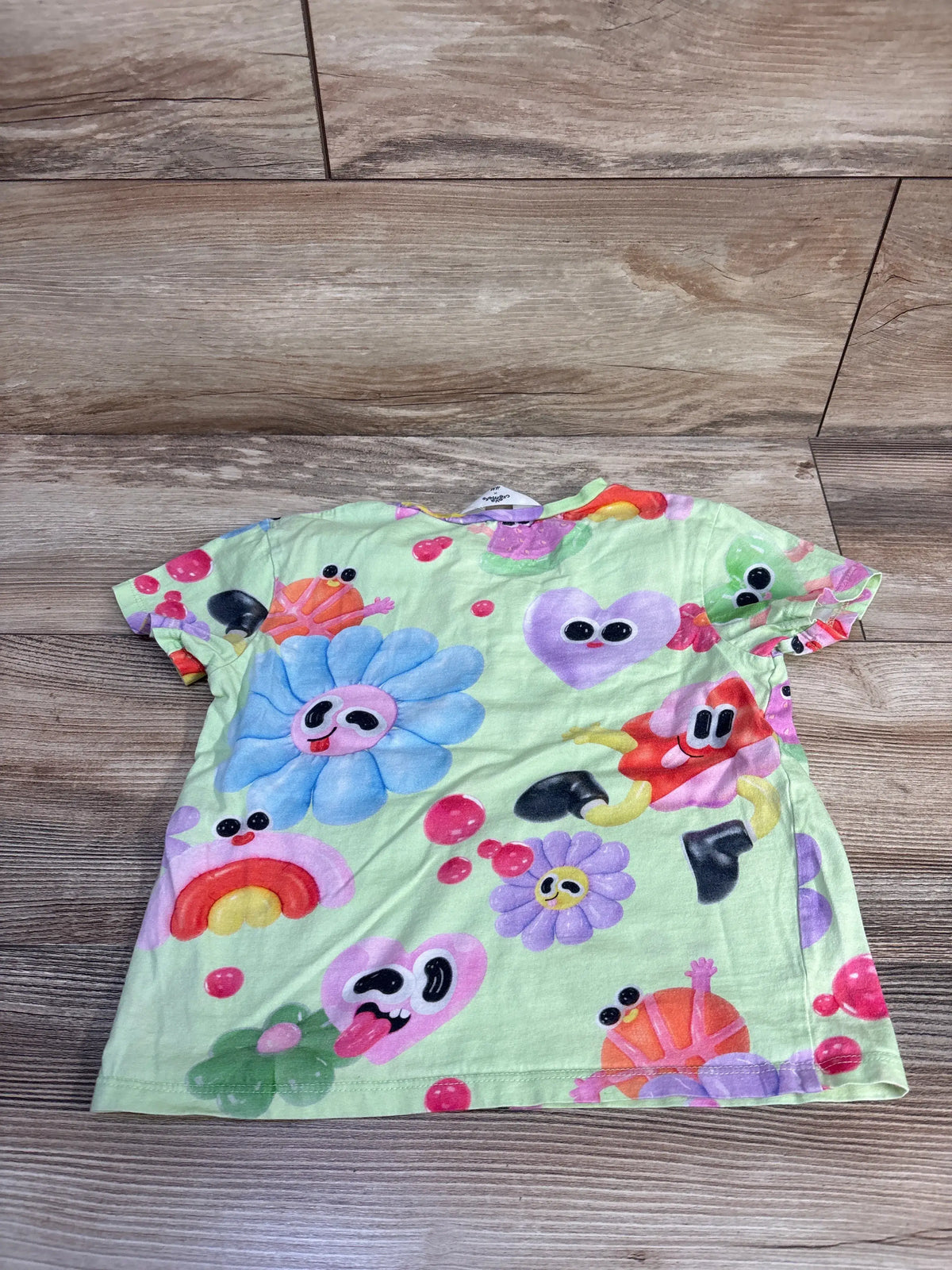 Front view of Eva Cremers x H&M green toddler shirt showcasing colorful cartoon and flower print on a wood floor